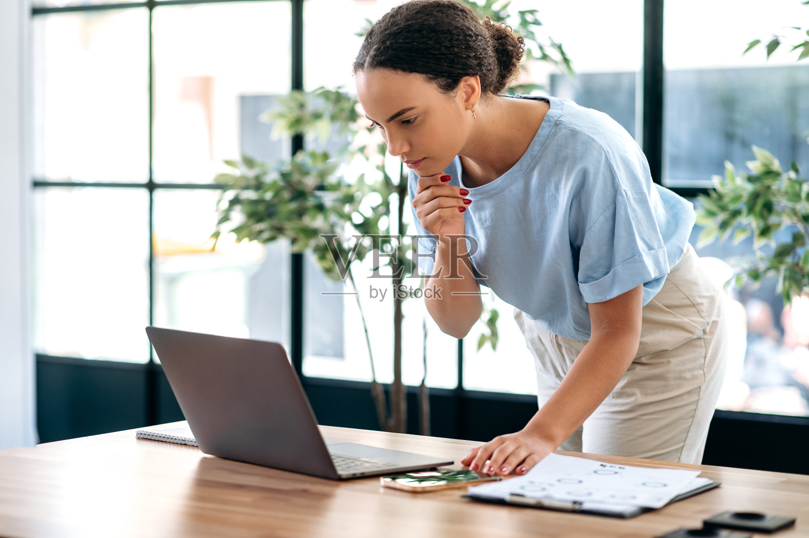 专注忙碌的巴西或西班牙女性员工，在工作文书工作项目中使用智能手机，站在现代化的办公室里。成功的混合种族商业妇女有手机通话照片摄影图片