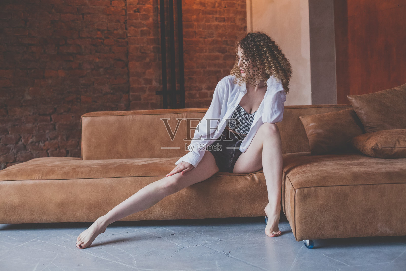 穿着白衬衫和短裤的卷发美女坐在棕色沙发上照片摄影图片