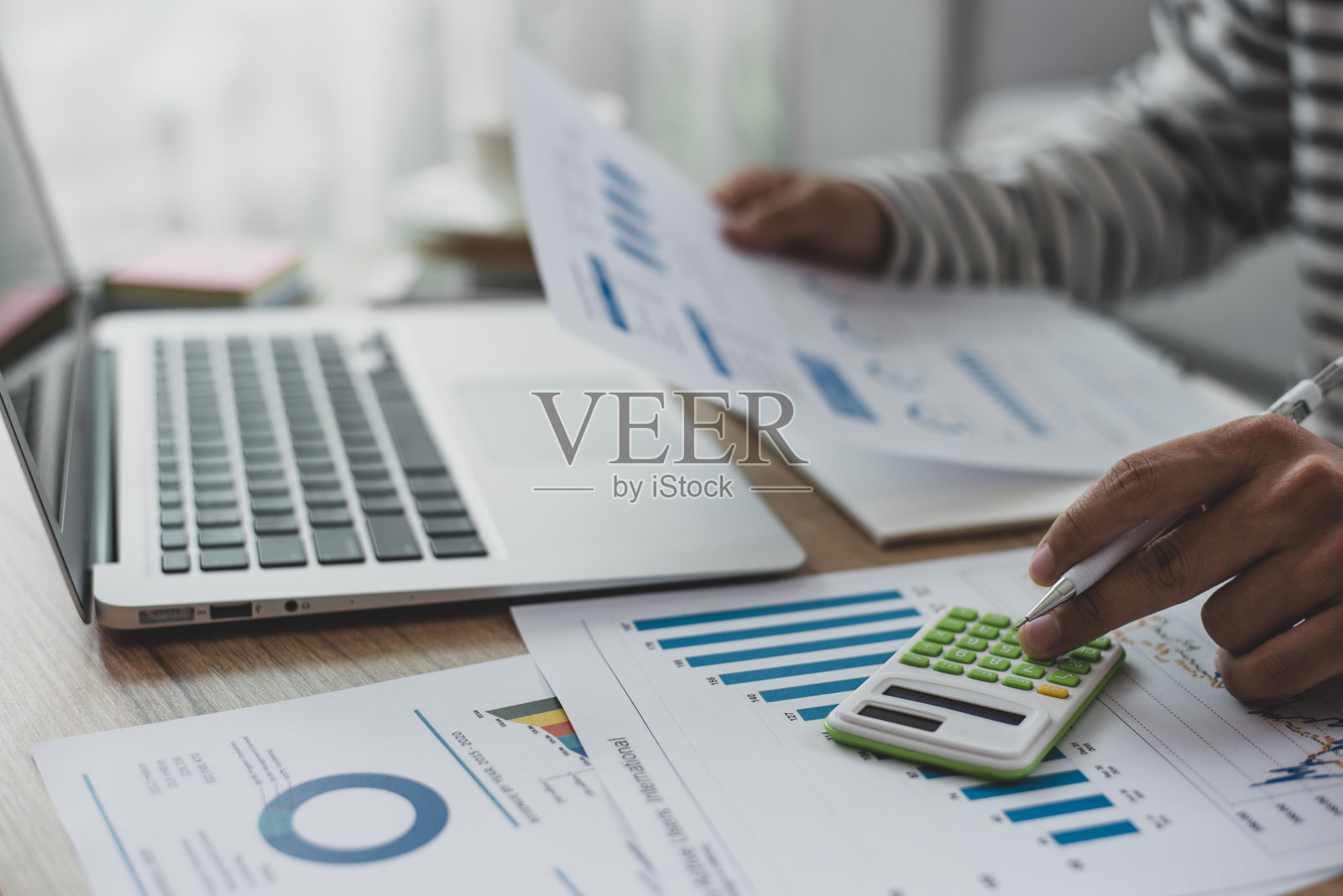 一名会计使用计算器和笔记本电脑，拿着一支笔，在笔记本上做笔记，检查财务报告文件躺在他的家庭办公室的桌子上。照片摄影图片