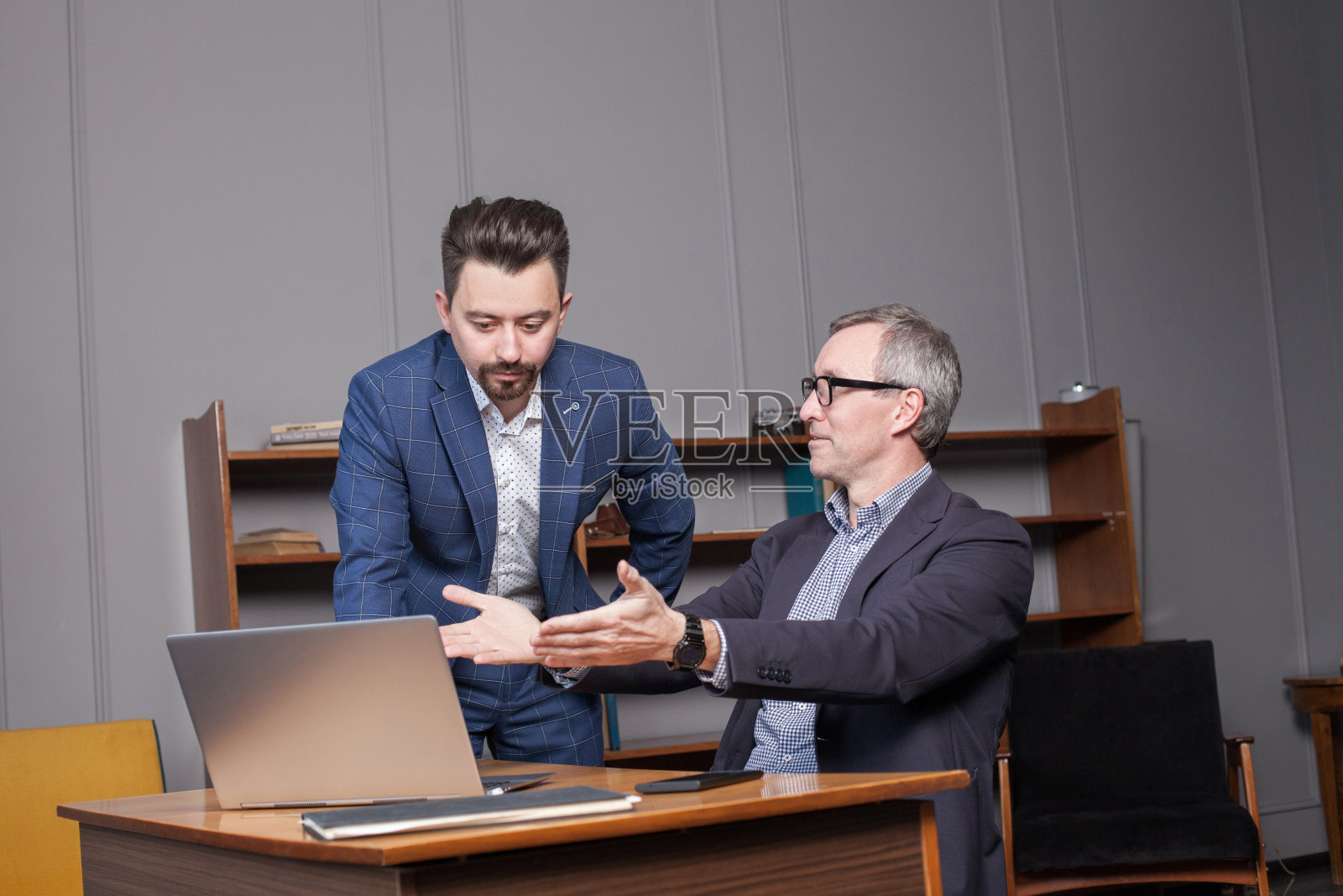 穿着蓝色西装、戴着眼镜的成熟商人正在办公室的笔记本电脑前与商业伙伴谈论工作照片摄影图片