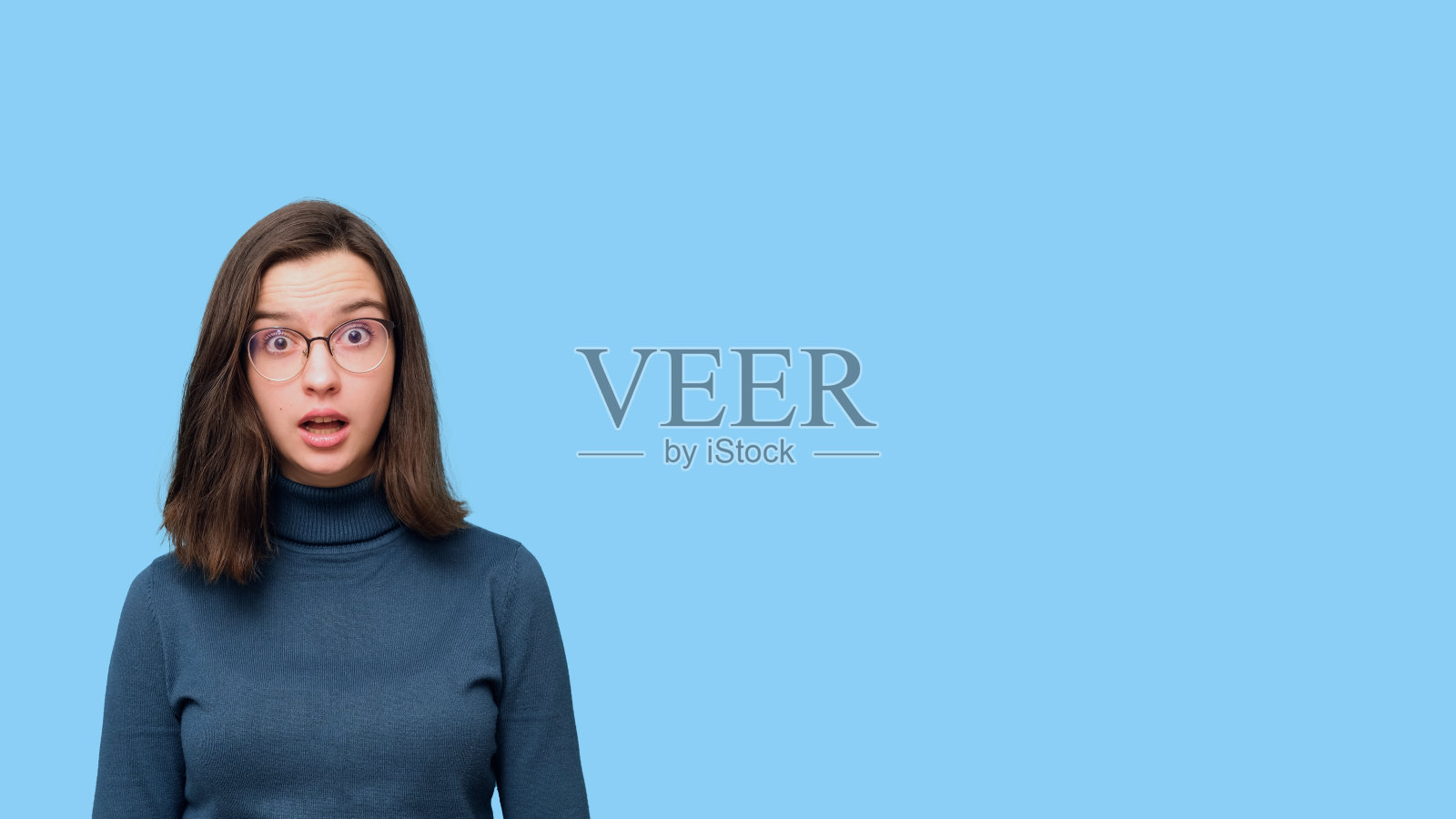 惊讶的女学生张着嘴，戴着眼镜和毛衣的肖像，蓝色背景隔离照片摄影图片