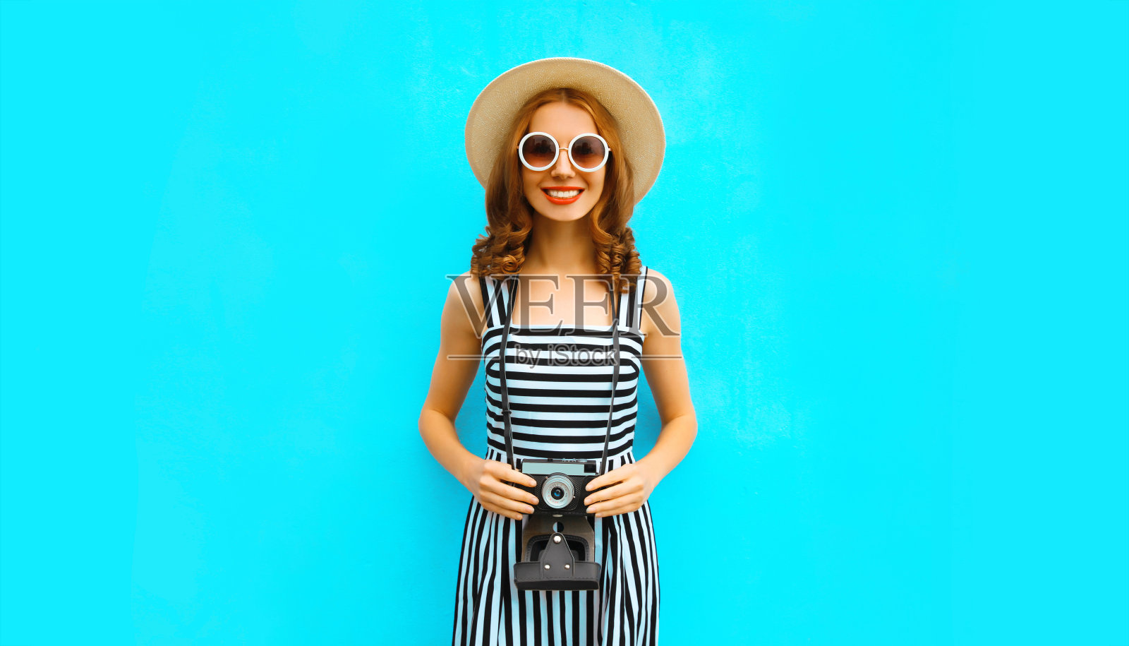 夏天的肖像快乐微笑的年轻女子摄影师与老式电影相机戴草帽，裙子，太阳镜在蓝色的背景照片摄影图片