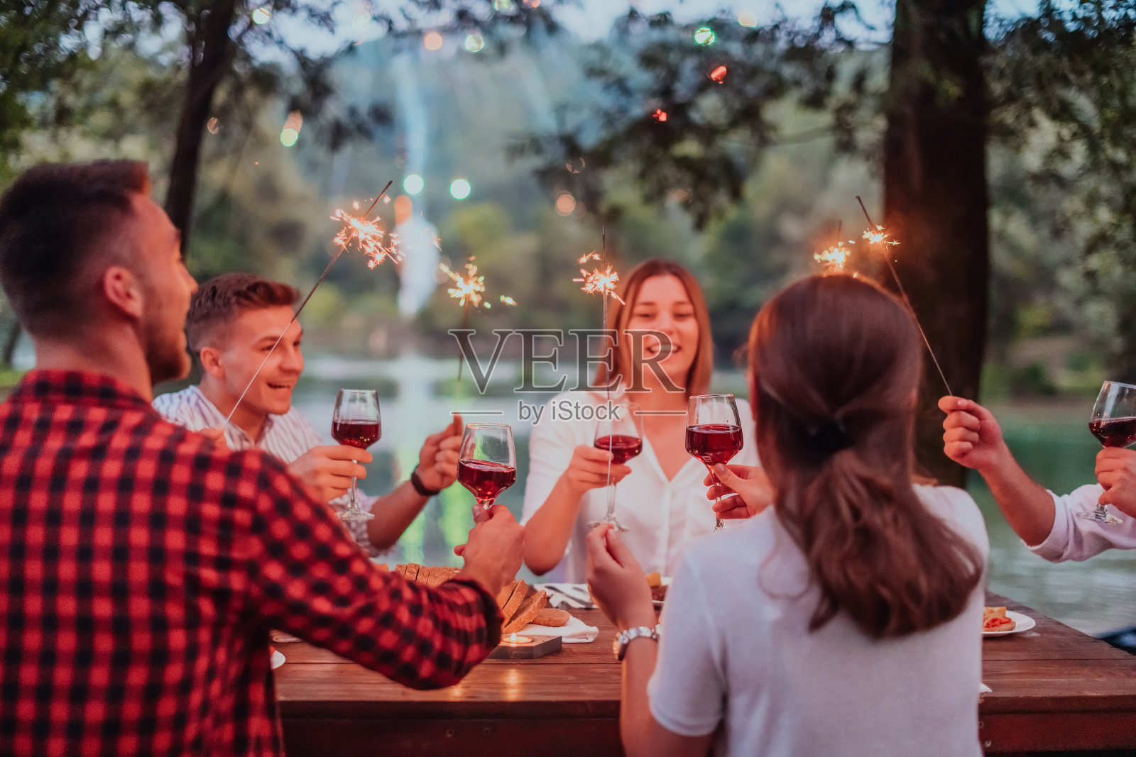 一群快乐的朋友在大自然美丽的夏夜，在河边的户外野餐法式晚餐派对上，用洒水器和红酒庆祝假期照片摄影图片