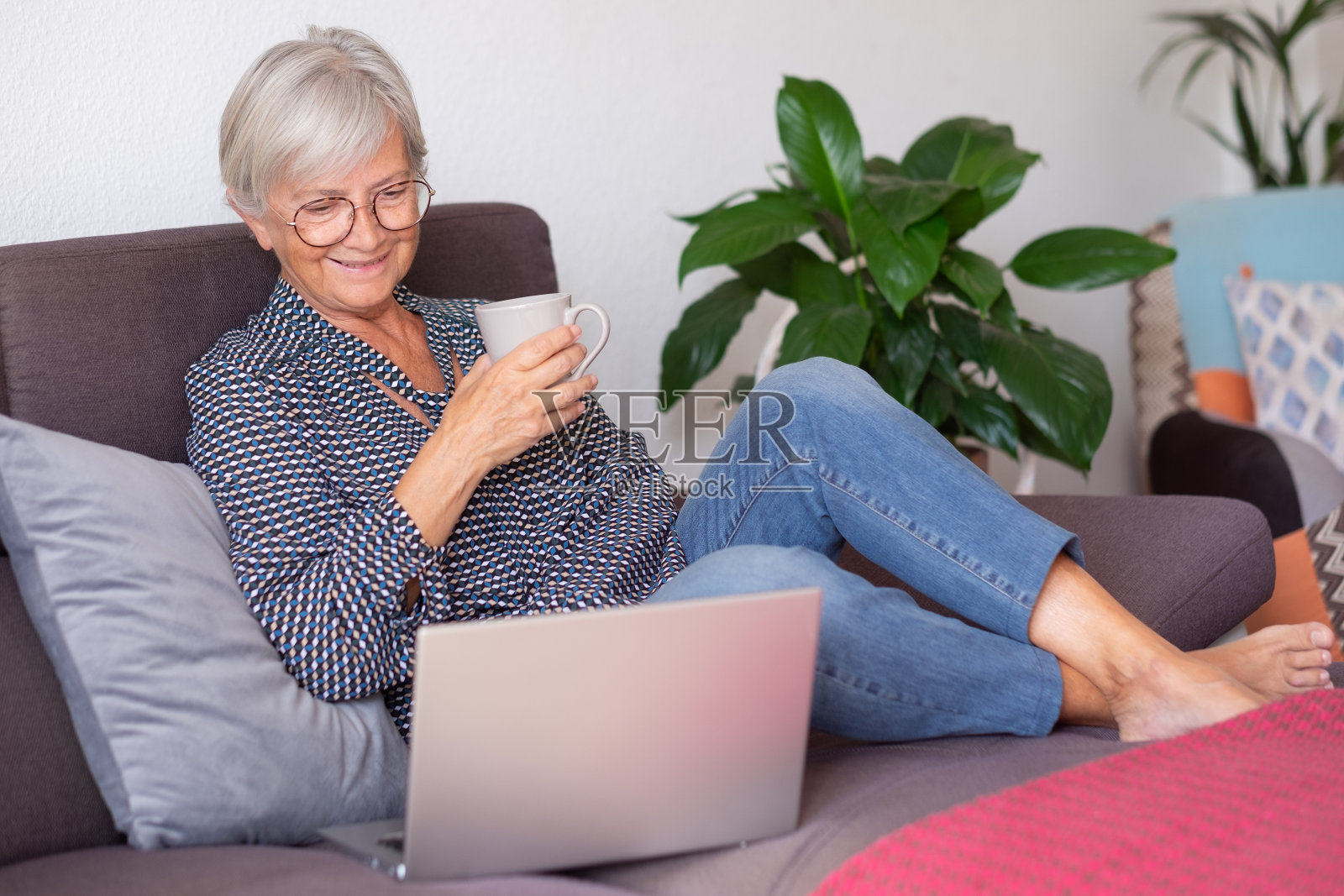 微笑的白人老太太坐在家里的沙发上，拿着咖啡杯，在笔记本电脑上浏览。照片摄影图片