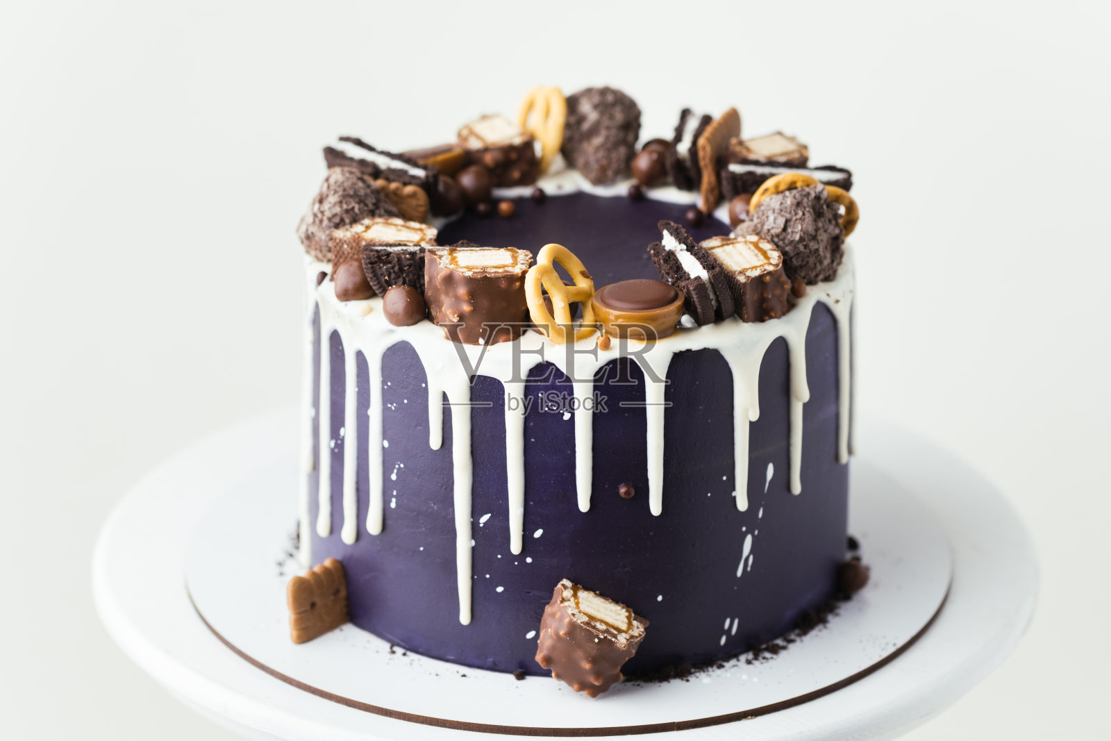 生日蛋糕，深蓝色奶油奶酪糖霜，白色背景上有白巧克力滴。白色的架子上有巧克力糖果和饼干装饰的自制蛋糕照片摄影图片