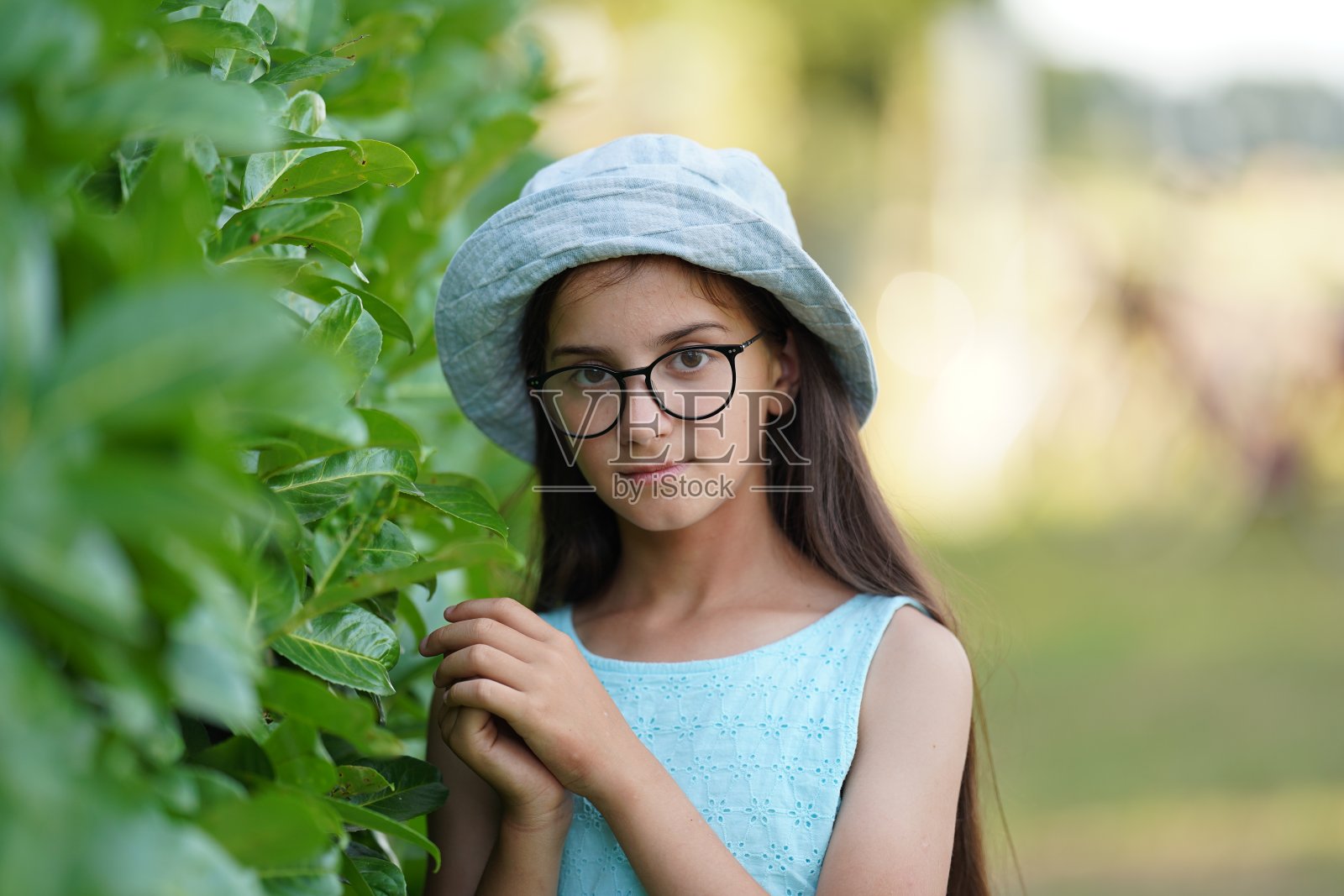 一个可爱的女孩留着长长的黑发，穿着浅色的裙子，穿着巴拿马大衣，戴着眼镜，在绿色的灌木丛旁摆姿势。有选择性的重点照片摄影图片