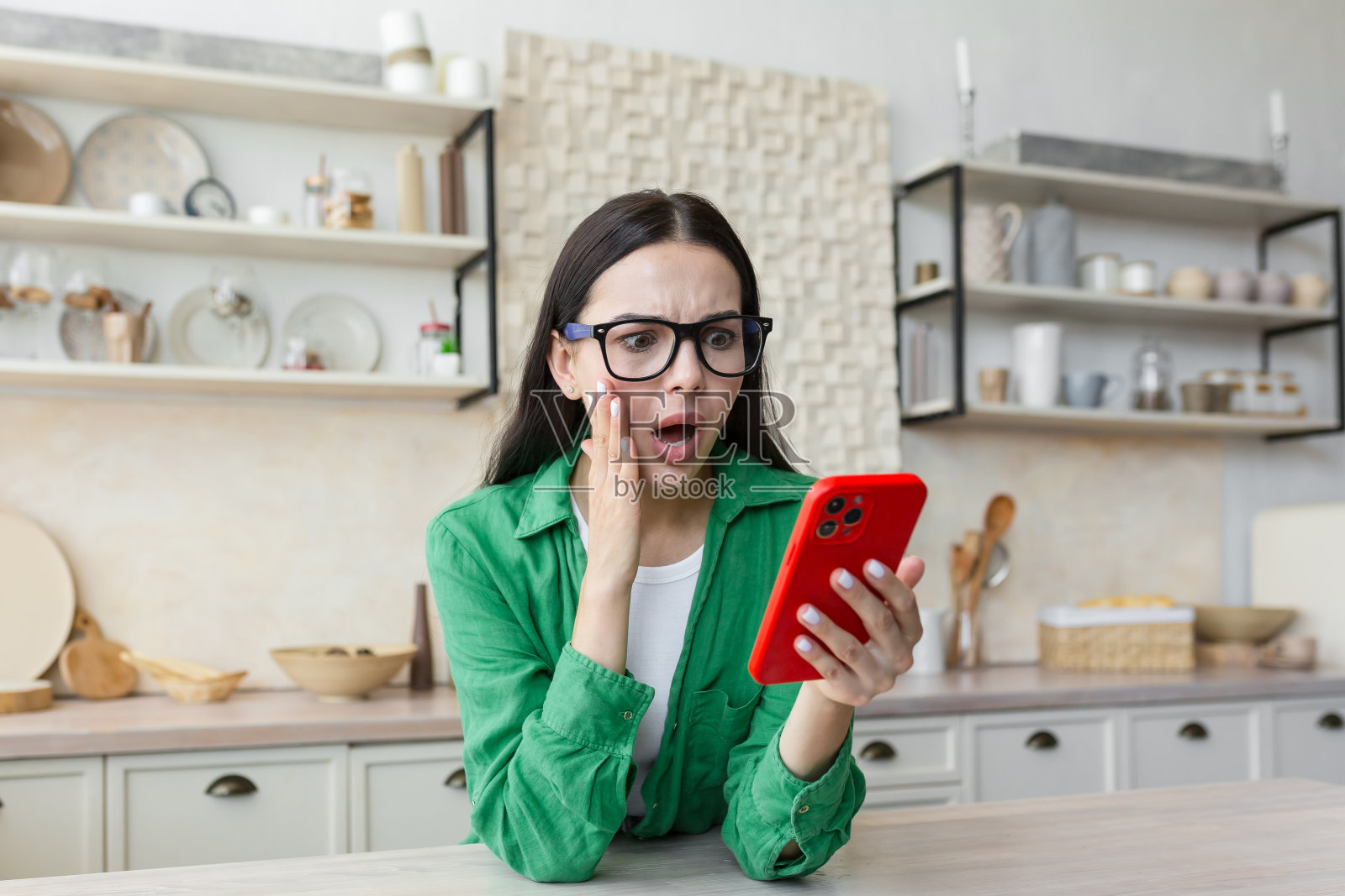 一个年轻的女人担心而震惊地站在家里的厨房里，手里拿着手机照片摄影图片