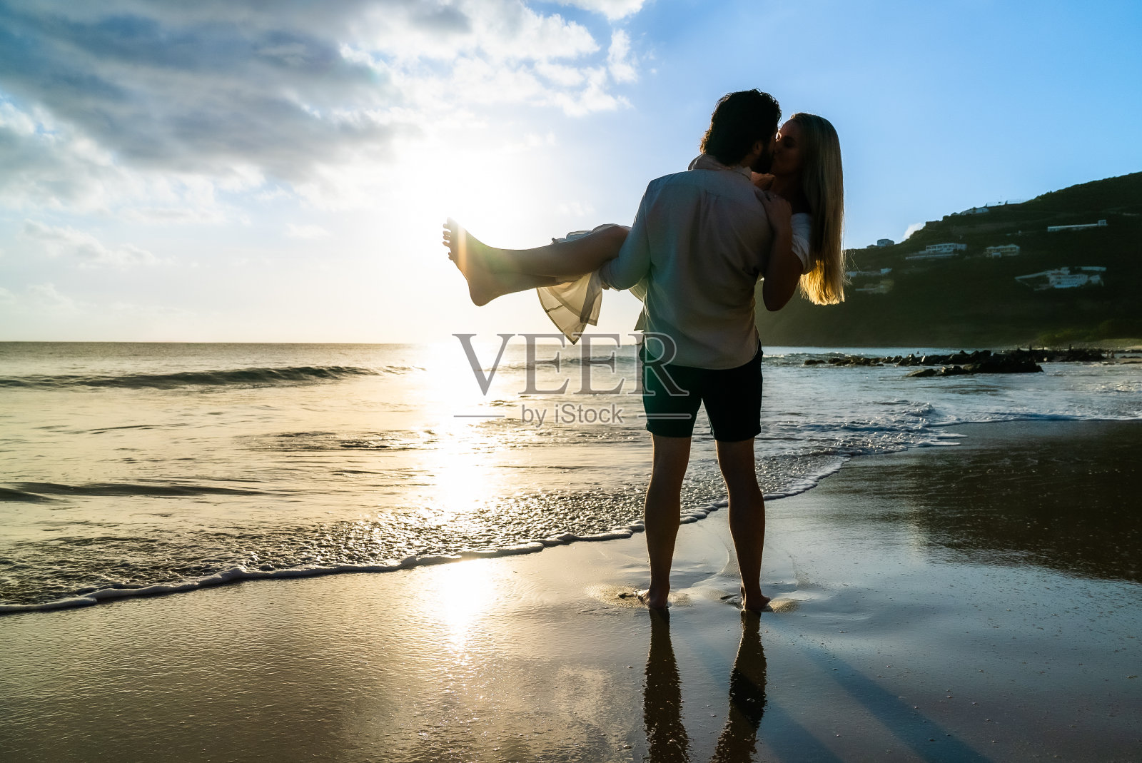 年轻的情侣在热带岛屿的海滩上亲吻，年轻的男子抱着他的女朋友在日落的天空中照片摄影图片