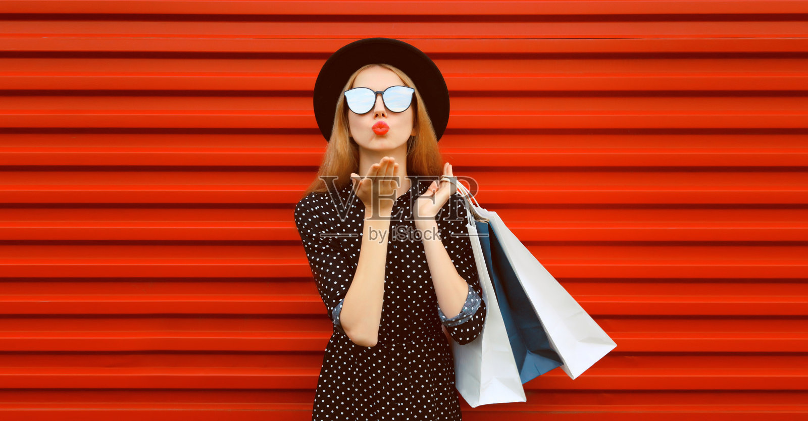 美丽的年轻女子的肖像购物袋吹她的嘴唇发送空中吻戴黑色圆帽在红色的背景照片摄影图片