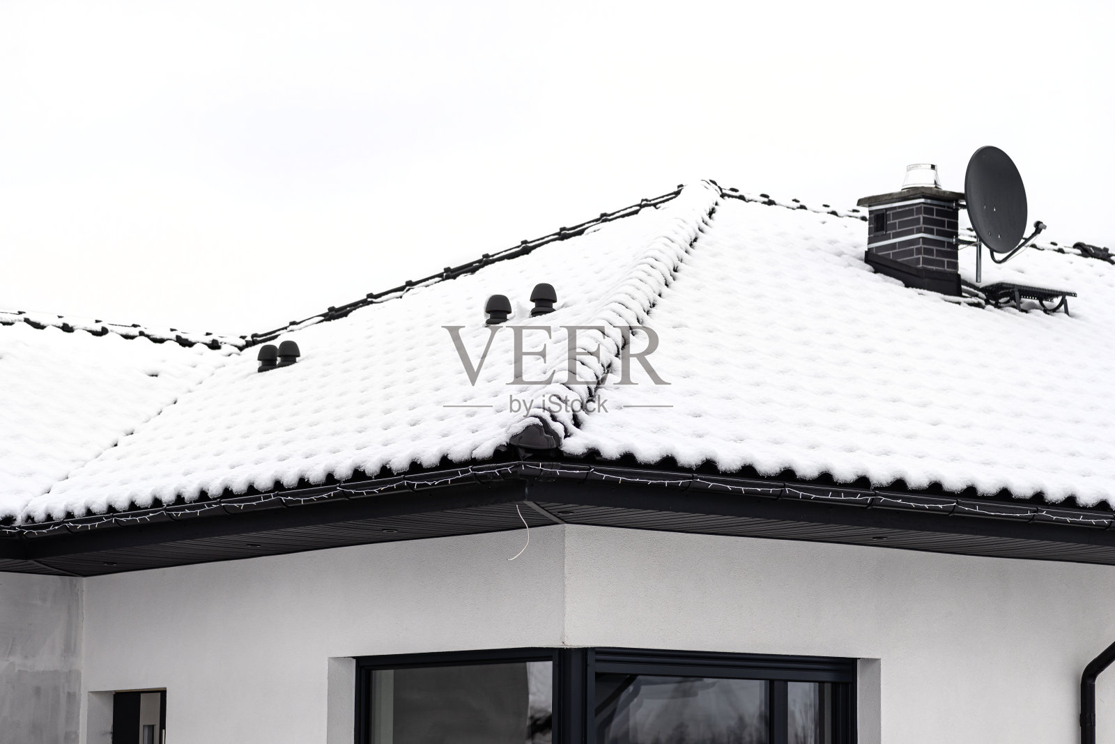 在多云的天空下，独栋住宅的屋顶覆盖着积雪，可以看到陶瓷通风壁炉和卫星天线。照片摄影图片
