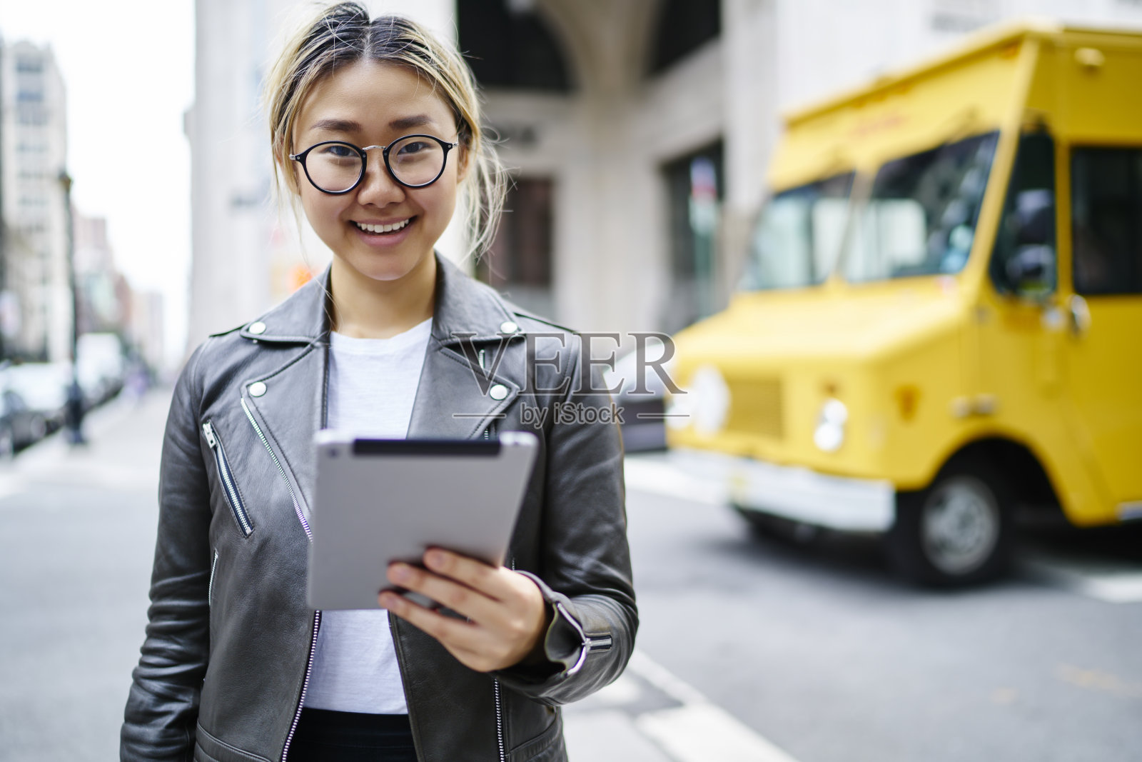 日本女游客在皮夹克手持现代数字平板电脑网络旅游网站和微笑在市中心的相机，快乐的千禧潮女孩的肖像触摸板照片摄影图片