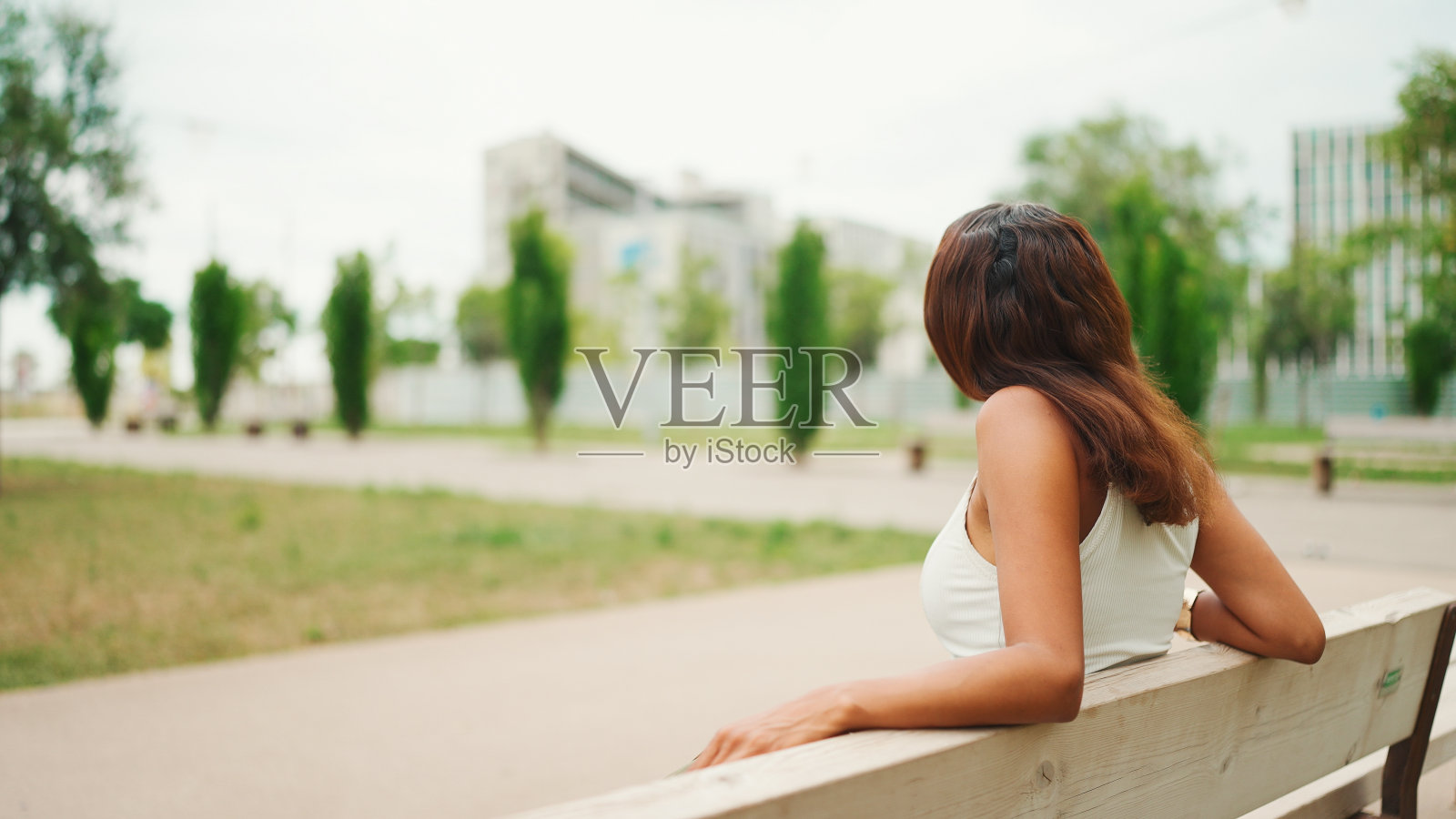 可爱的黝黑的女人，棕色的长发，穿着白色的上衣，坐在公园的长椅上放松地看着别处。生活方式概念。缓慢的运动。照片摄影图片