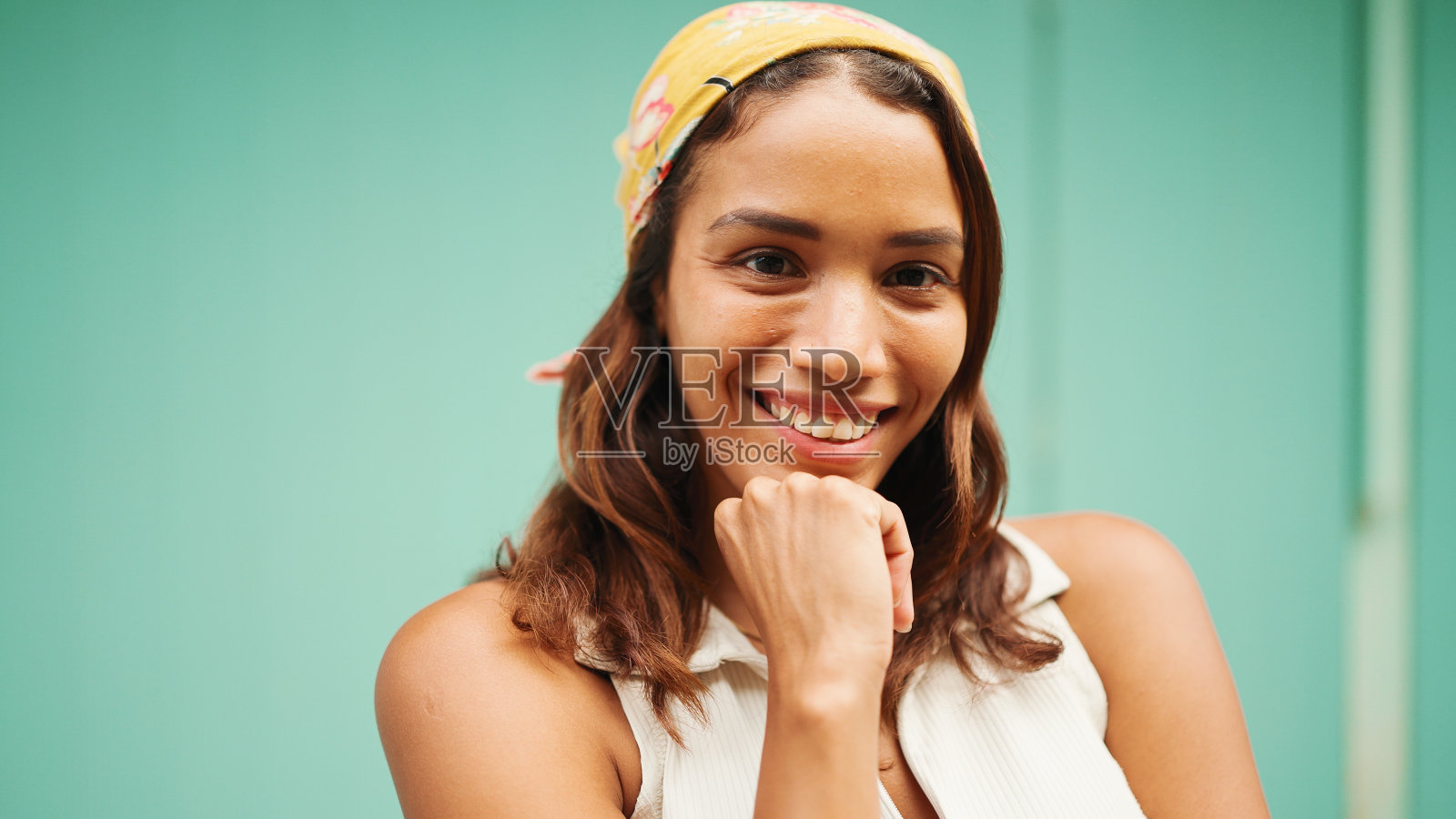 美丽的女孩，晒黑的皮肤，棕色的长发，头上戴着大手帕，在镜头前摆姿势。微笑的年轻女子把一根手指放在蓝色背景的脸颊上照片摄影图片