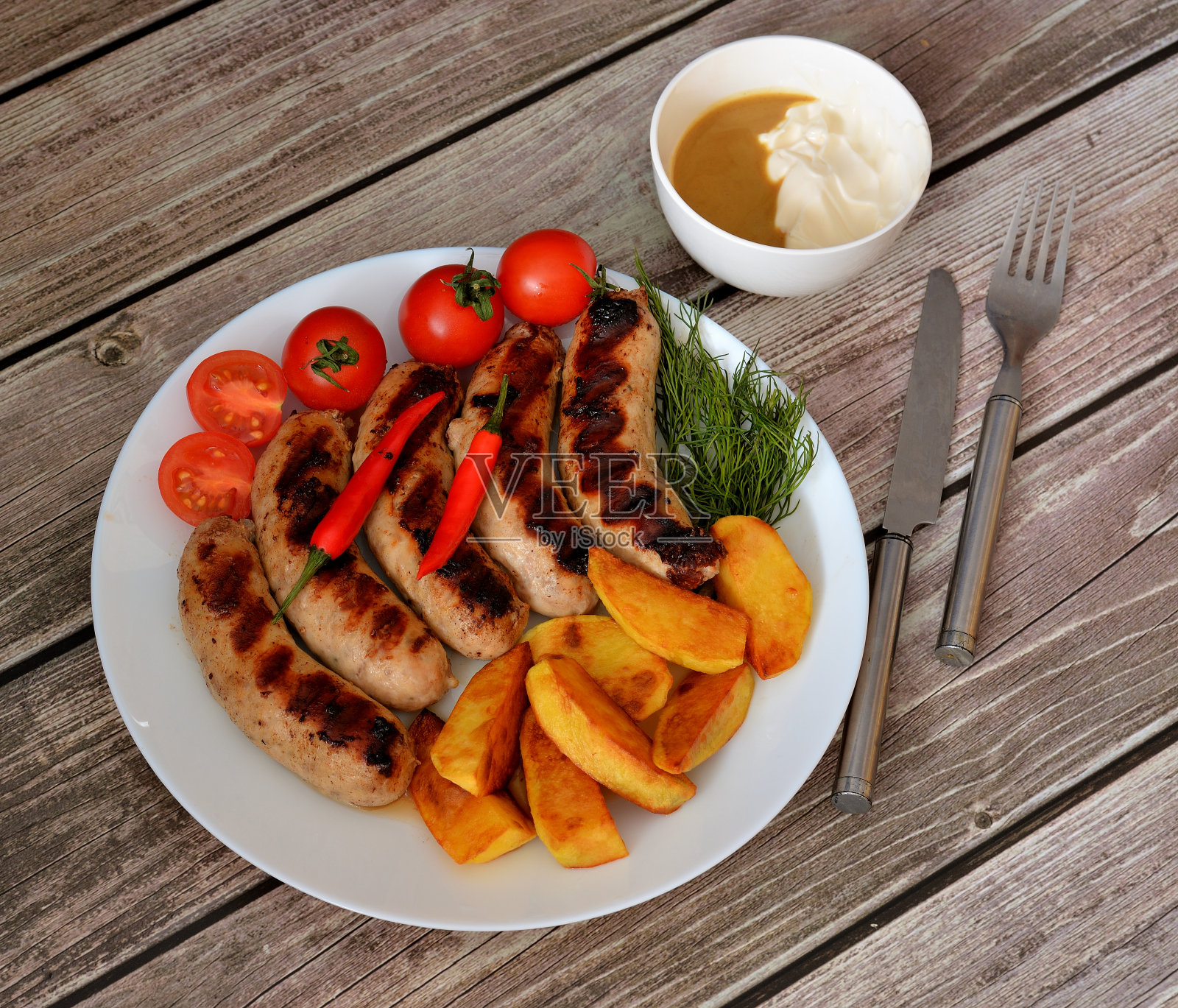 木桌上摆着一个盛着烤香肠、圣女果、香草、炸土豆和辣椒的盘子，还有一个盛着酱汁的碗。照片摄影图片