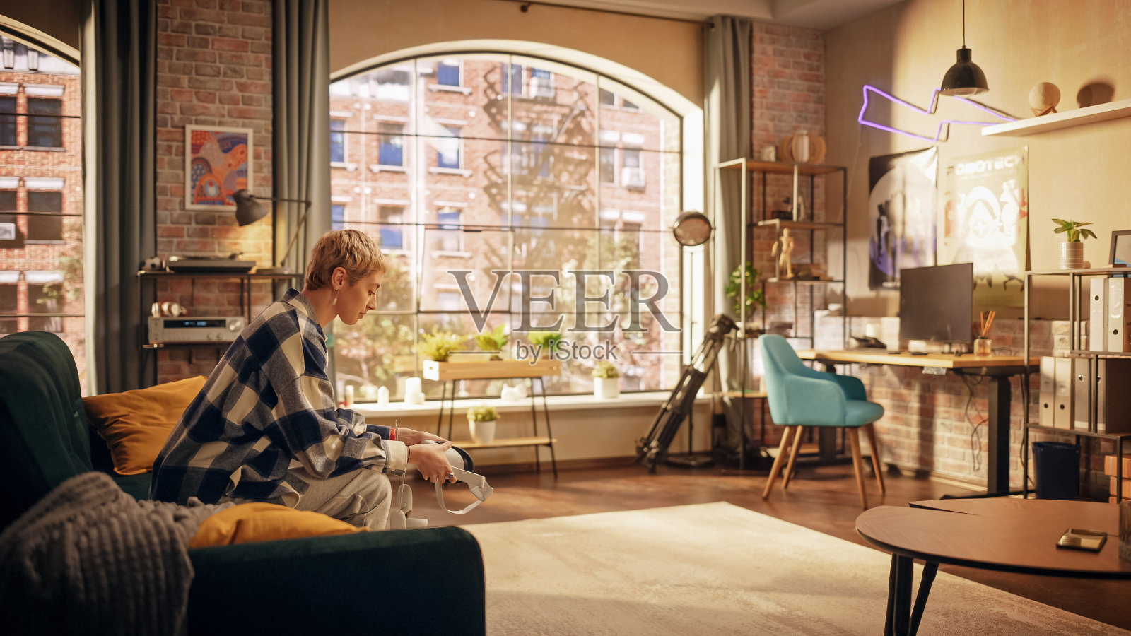 年轻的创意女性坐在阁楼时尚客厅的沙发上，手里拿着VR耳机。美丽的女人即将进入数字元宇宙世界与她的朋友们玩耍。照片摄影图片