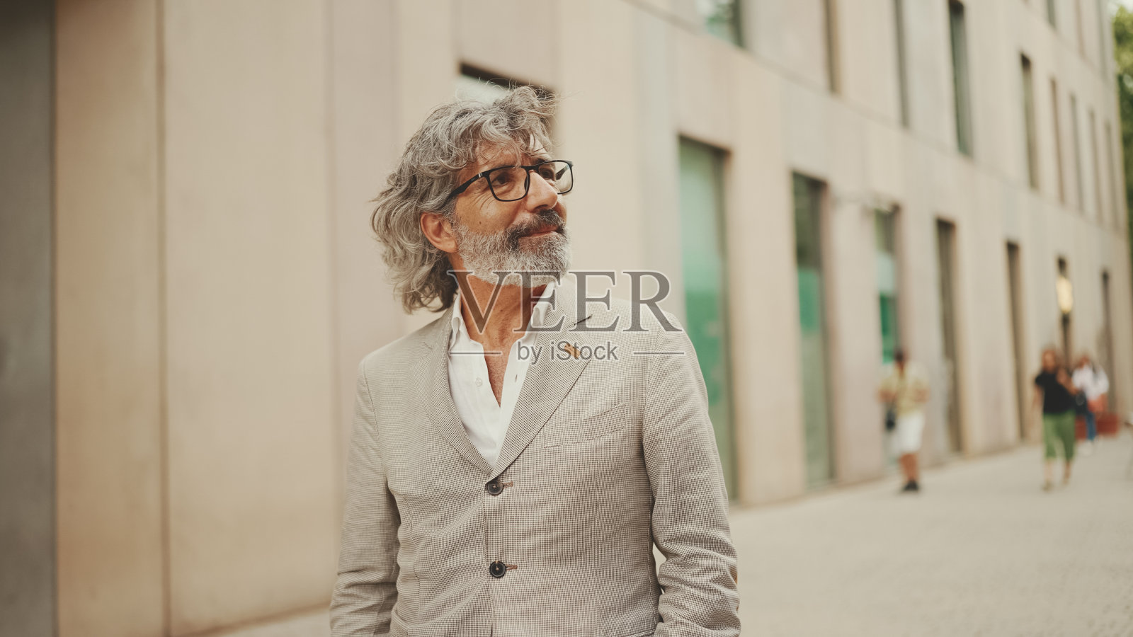 成熟的商人，留着胡子，戴着眼镜，穿着灰色夹克，走过现代化的建筑照片摄影图片