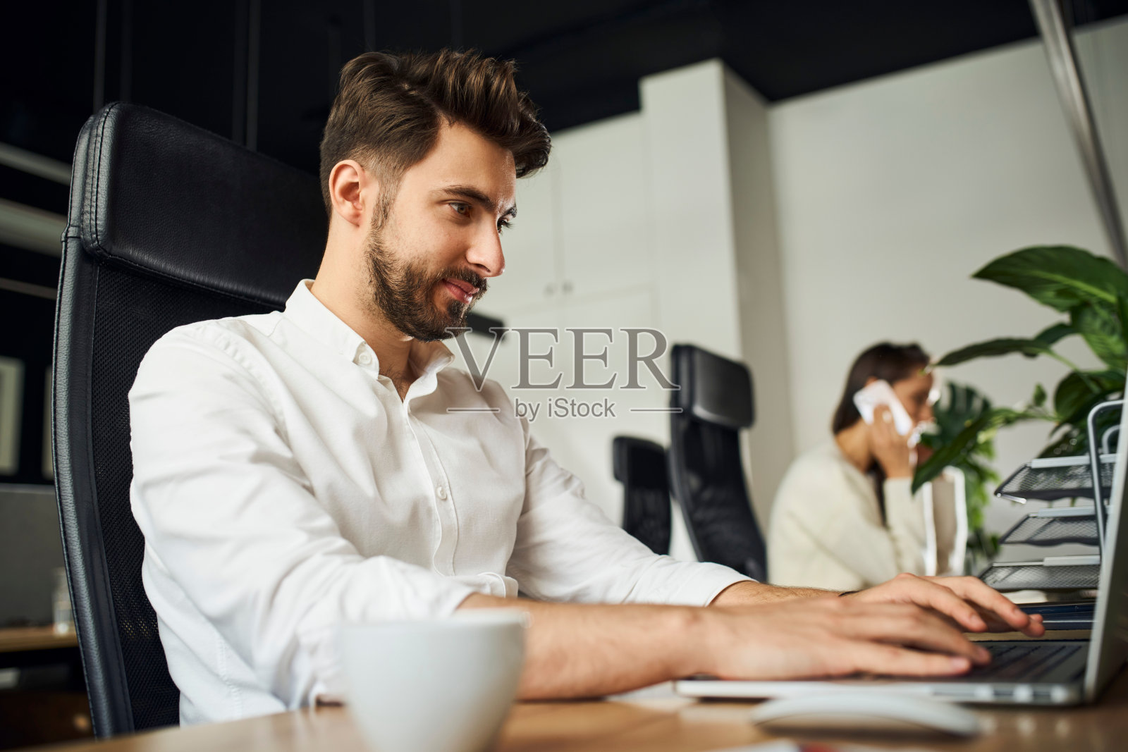 年轻商人的肖像工作在小办公室使用笔记本电脑与同事在后台照片摄影图片