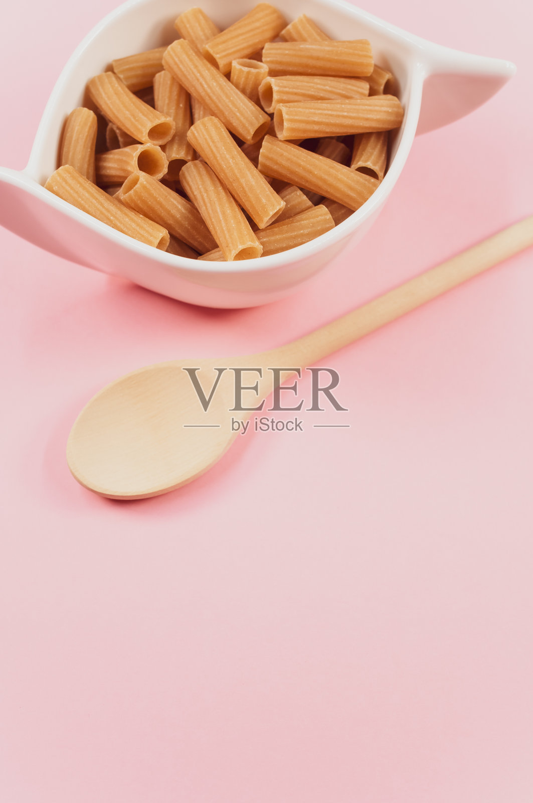 垂直拍摄一碗未煮熟的意大利面，一把木勺放在粉红色的表面上照片摄影图片