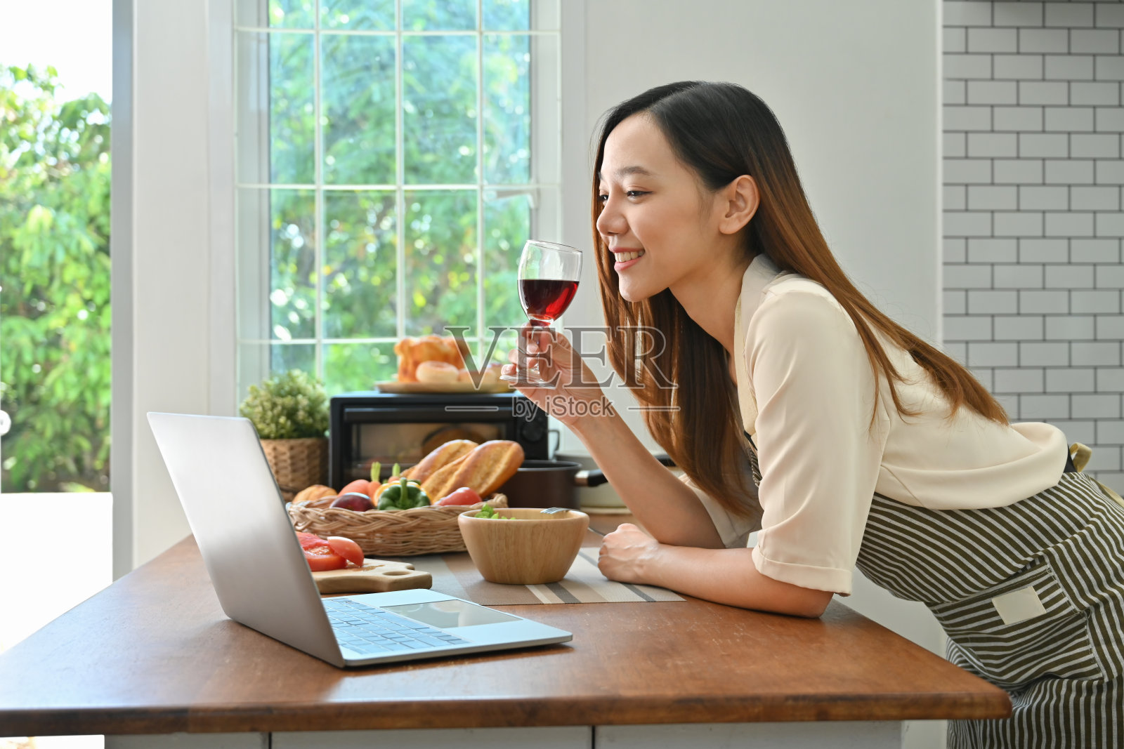 迷人的年轻女子喝着红酒，在厨房的笔记本电脑上通过视频通话与朋友在线聊天。照片摄影图片