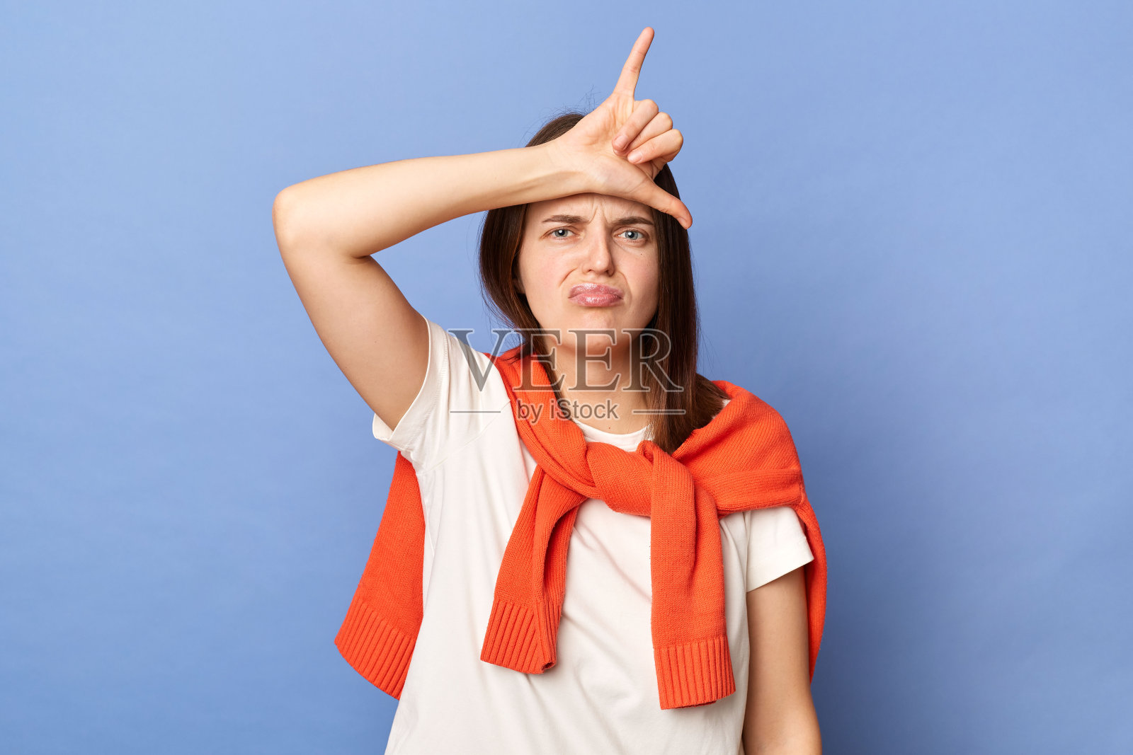 失望的悲伤不安的女人穿着白色t恤和系在肩膀上的毛衣站在蓝色背景上孤立的肖像，用手指显示手势，感觉更宽松。照片摄影图片