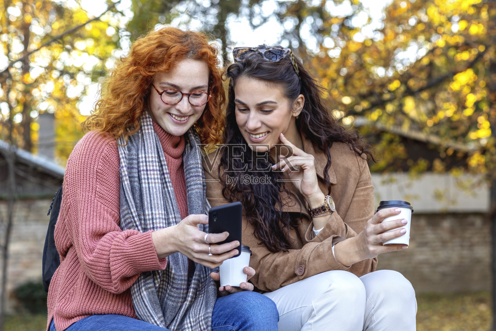 两个漂亮的女孩在校园附近的公园里聊天，手里拿着手机笑着玩照片摄影图片