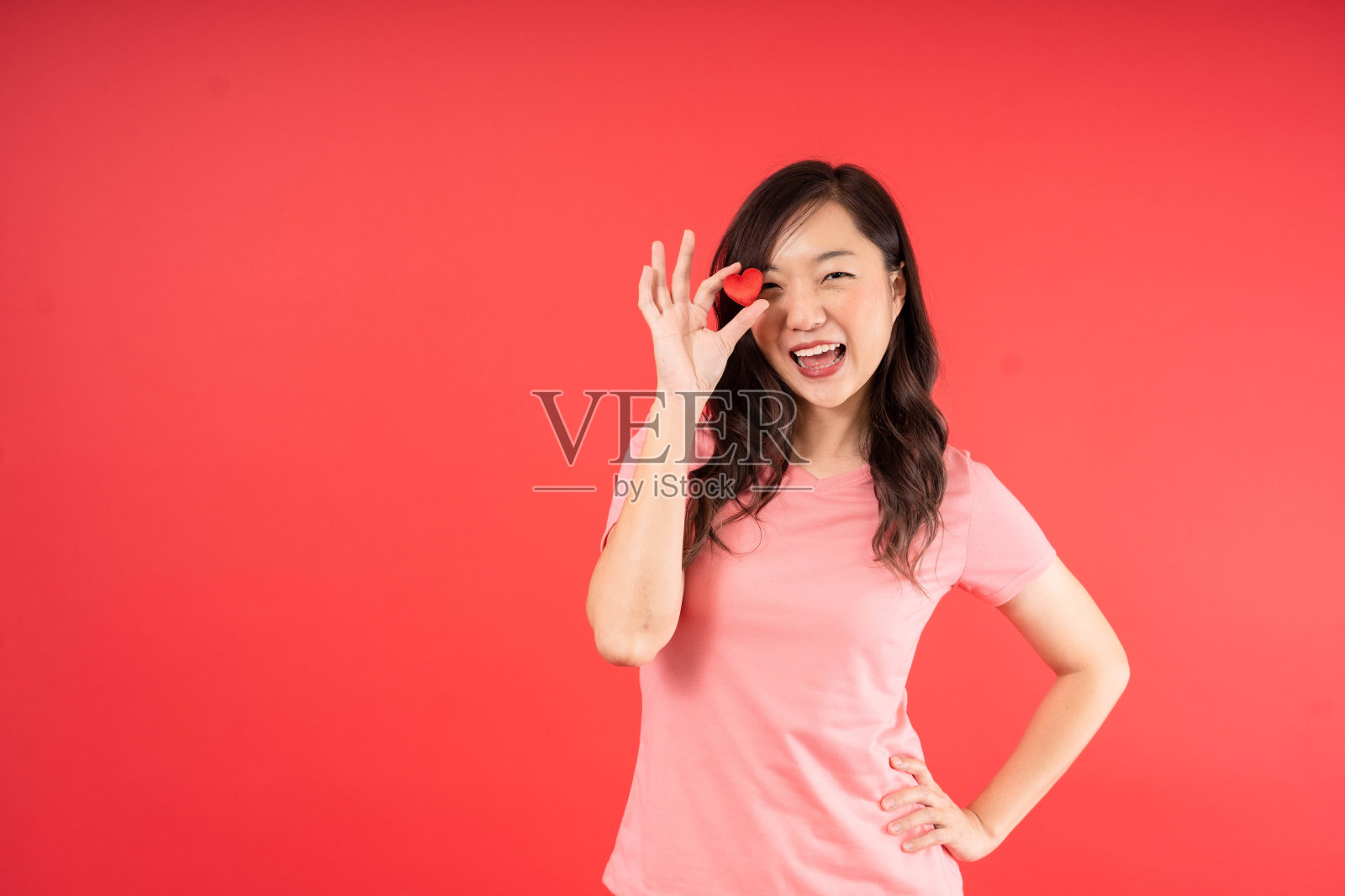 美丽的亚洲女人，粉红色的衬衫感到快乐，抱着小小的心孤立在红色的背景。情人节概念。照片摄影图片