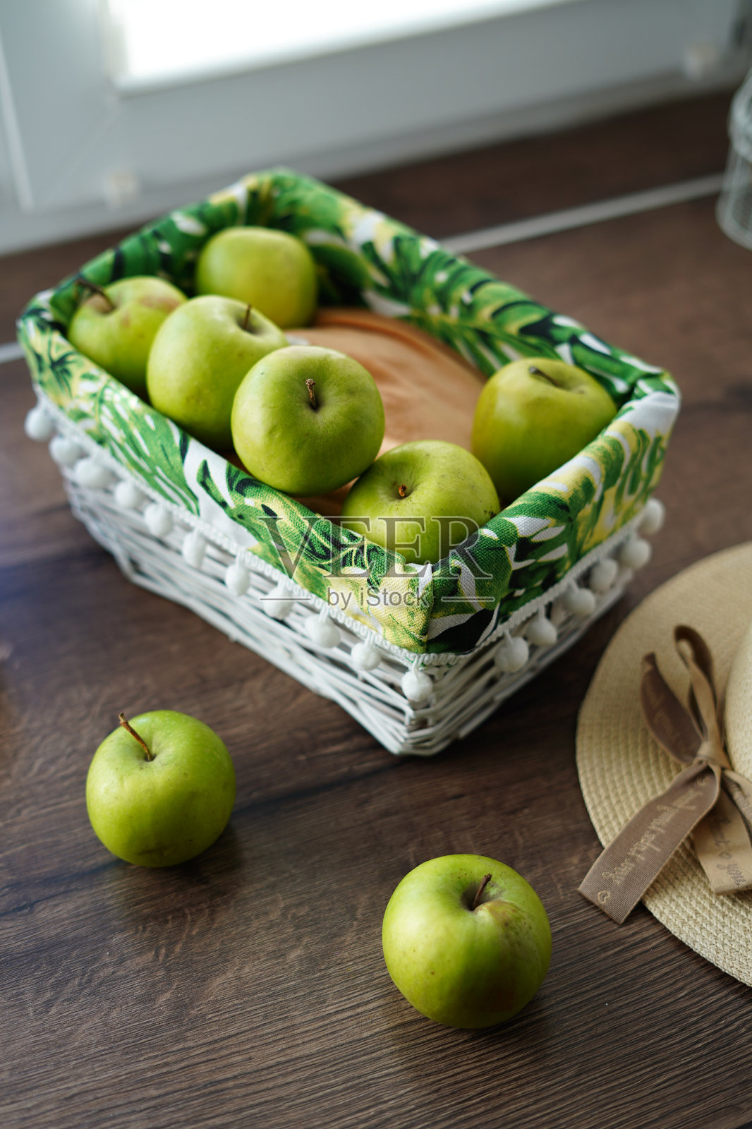 靠窗的木桌上放着一个白色的柳条篮子，里面放着绿色多汁的苹果。篮子旁边放着一顶米色帽子和一个胡桃木篮子。阳光明媚的一天。漂亮的背景照片摄影图片