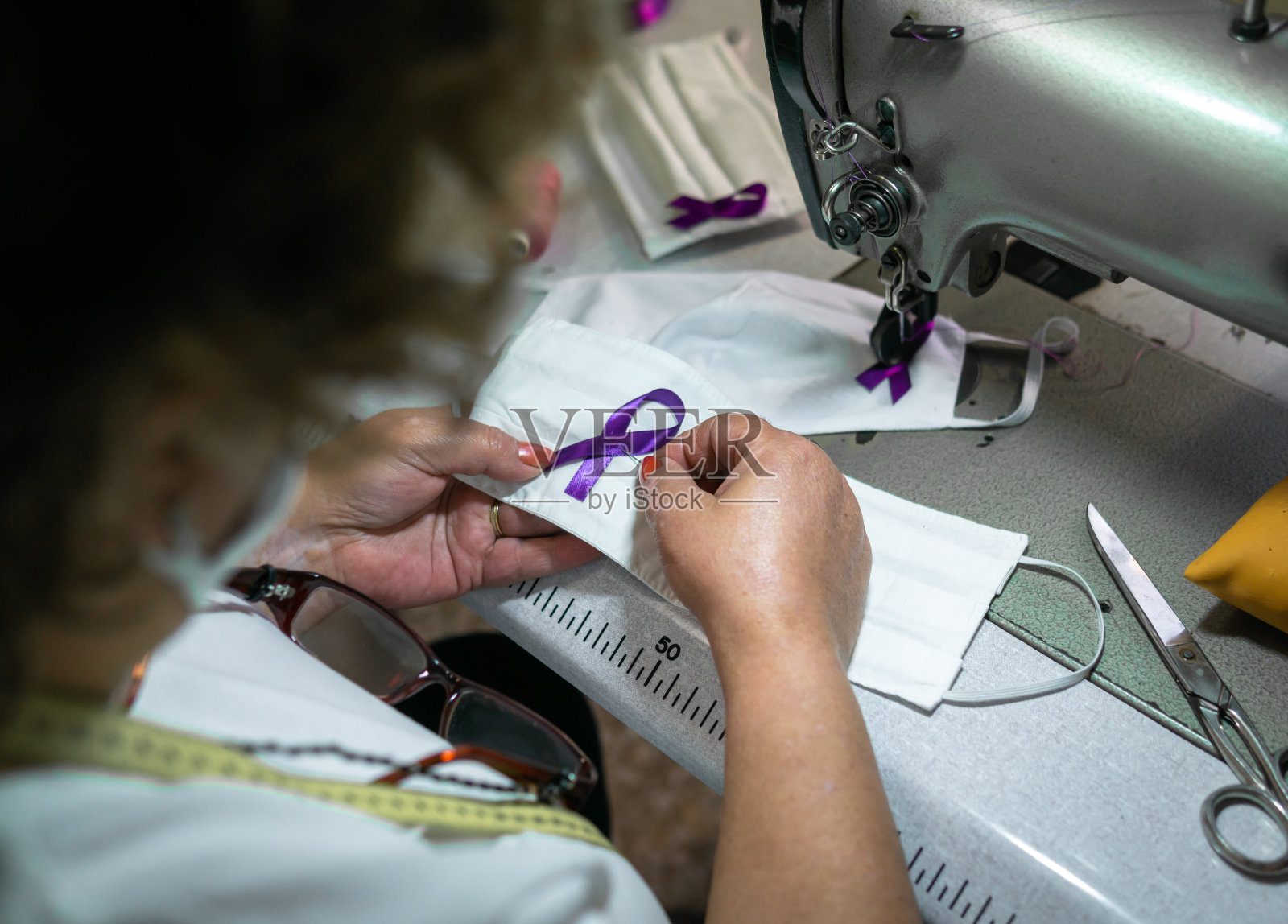 一名成年妇女正在车间为口罩缝制紫色丝带——COVID-19意识照片摄影图片