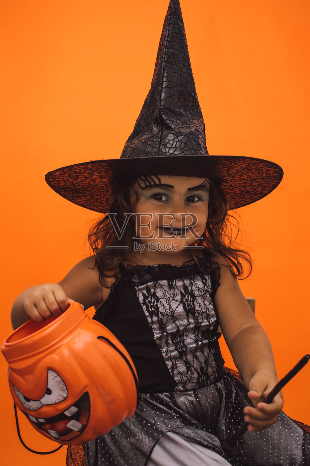 一个孩子穿着女巫服装，手里拿着一个南瓜，橙色背景的垂直镜头照片摄影图片