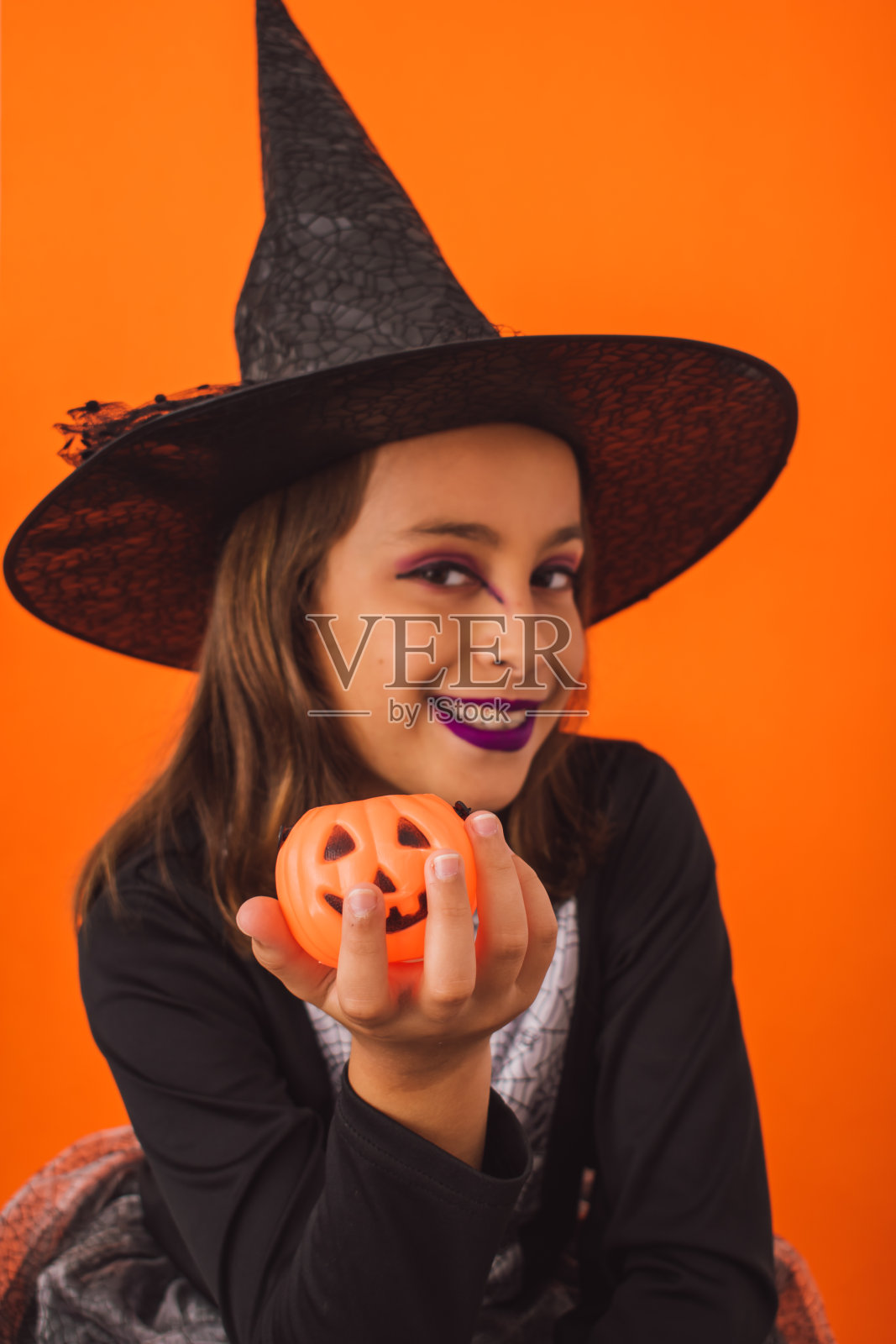 一个孩子穿着女巫服装，手里拿着一个南瓜，橙色背景的垂直镜头照片摄影图片