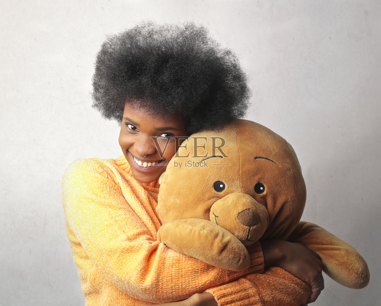 年轻的黑人女性，短发，穿着黄色毛衣，抱着一只泰迪熊靠在墙上照片摄影图片