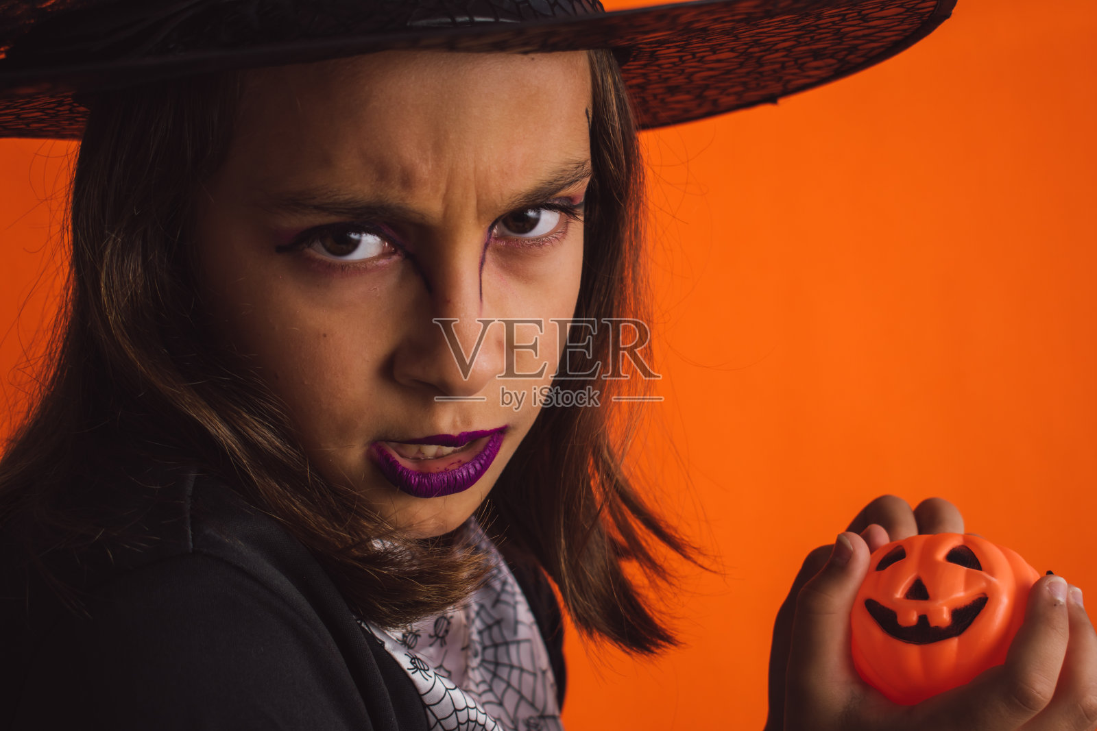 小女巫拿着一个万圣节南瓜，一个愤怒的手势和脸在一个橙色的背景照片摄影图片
