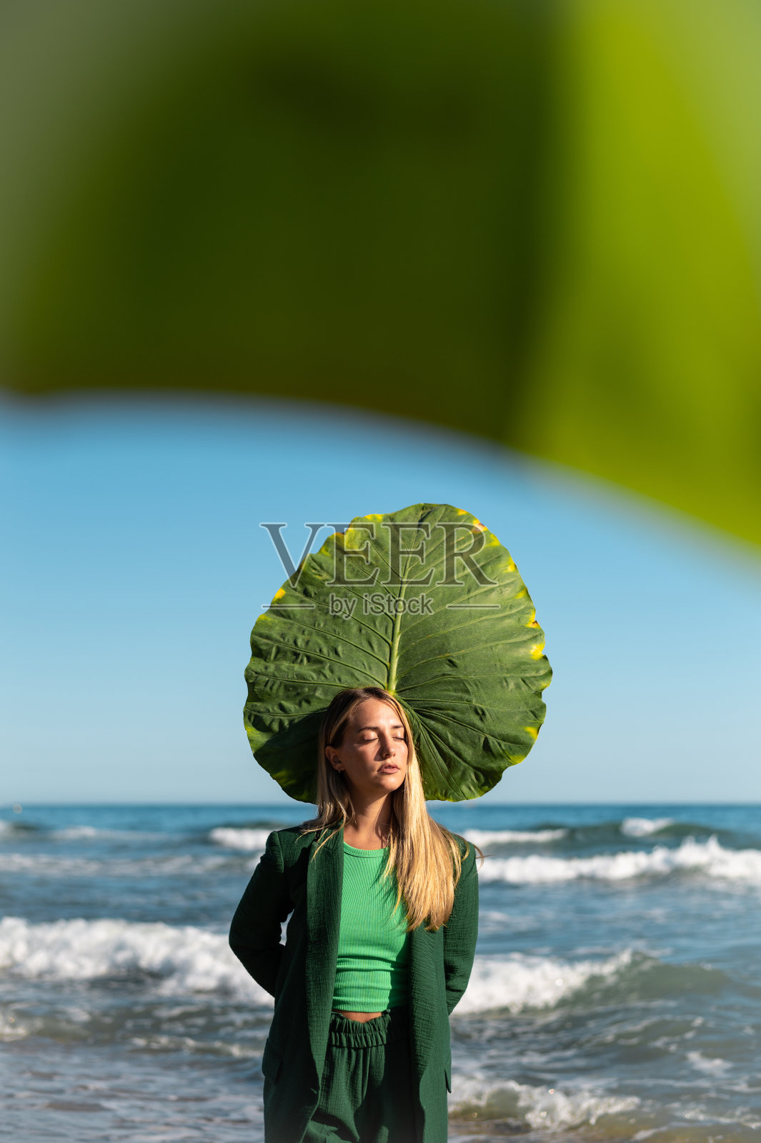 一个金发碧眼的西班牙女孩站在海滩上，手里拿着一片巨大的绿叶照片摄影图片