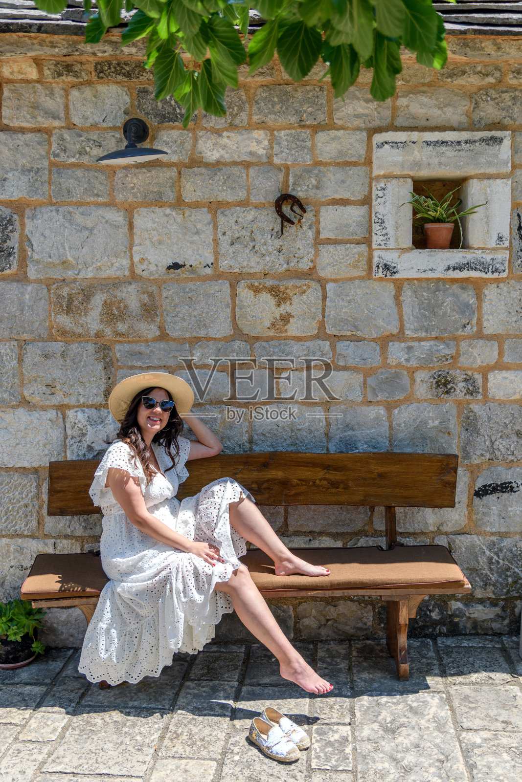 美丽快乐的年轻女子穿着白色连衣裙，坐在老石屋前的长凳上照片摄影图片