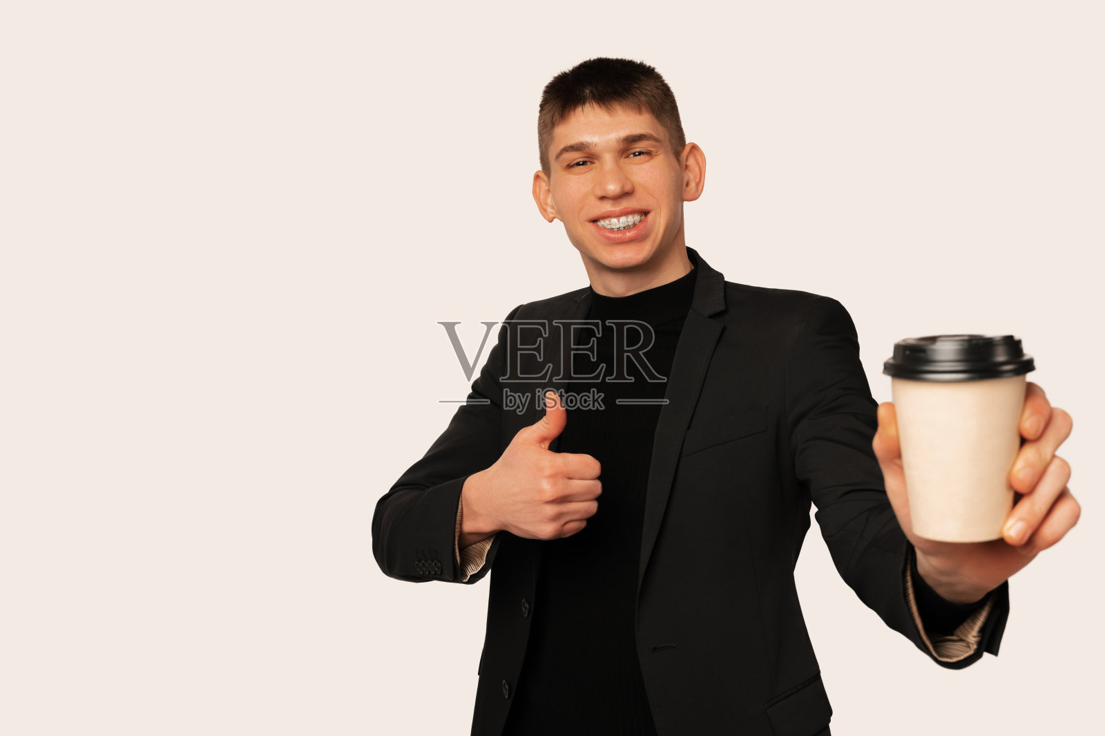 穿着夹克的英俊的年轻人拿着一个外卖杯，竖起大拇指。照片摄影图片