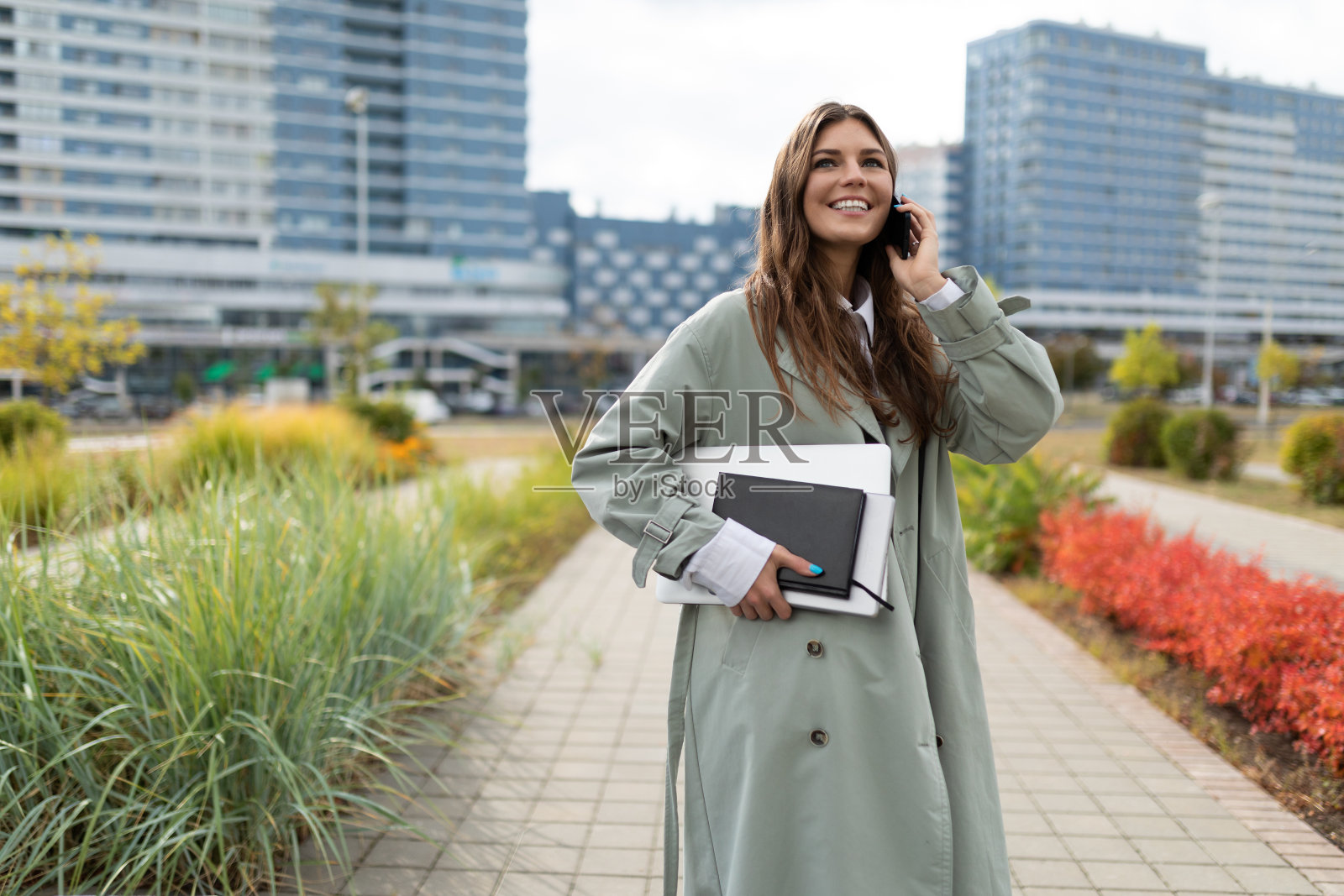 年轻的女学生，胳膊下夹着一台笔记本电脑，在城市的背景下打电话照片摄影图片