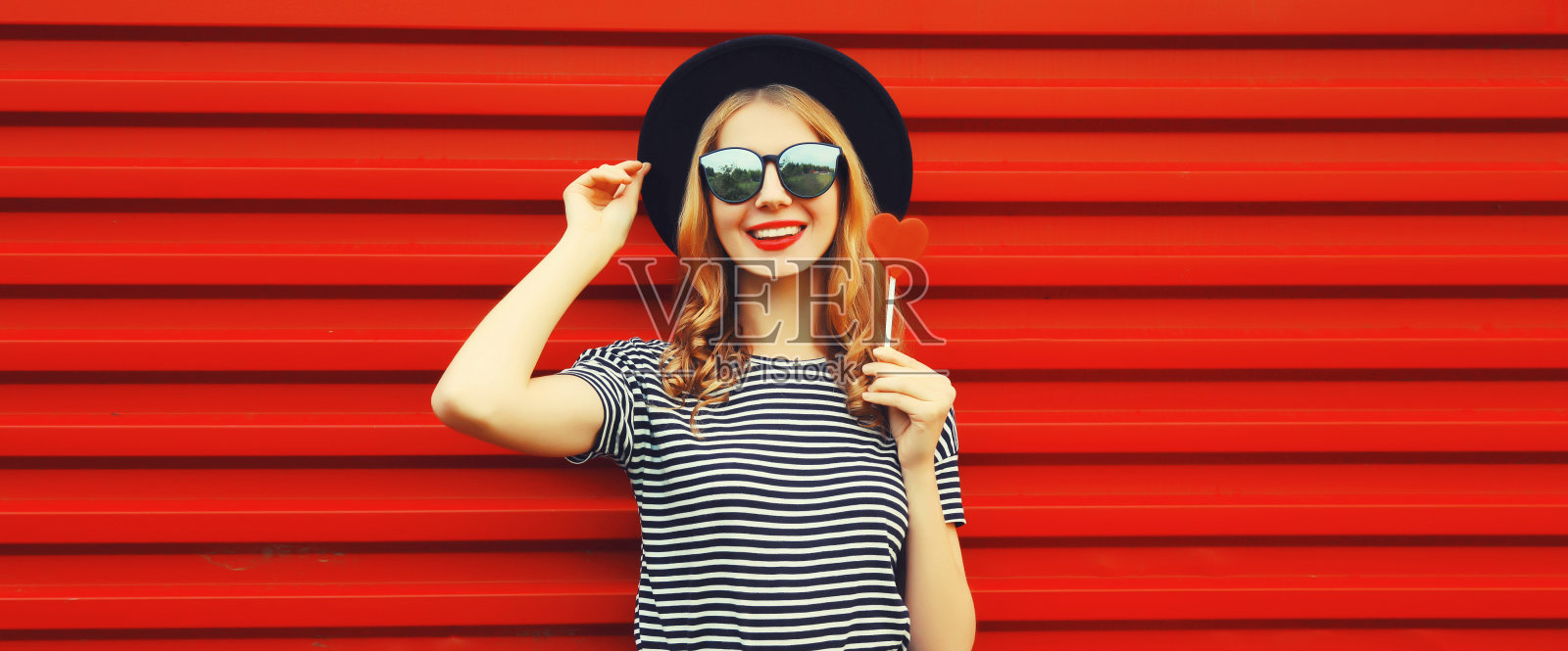 美丽的年轻女子肖像与红色心形棒棒糖戴太阳镜，黑色圆形帽子的背景照片摄影图片