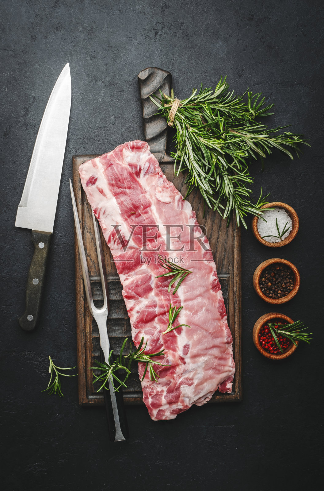 黑色厨房餐桌背景，俯视图，配上迷迭香和香料的生排骨，肉叉和厨师刀放在乡村木砧板上准备烹饪照片摄影图片