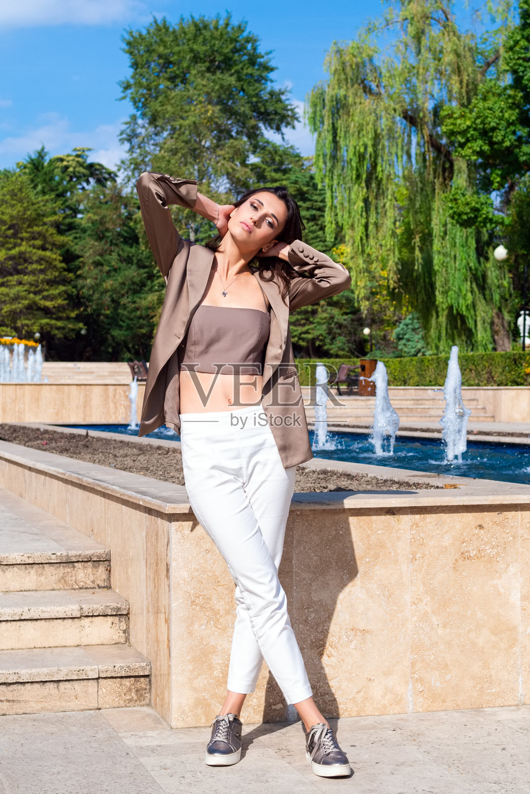 迷人的黑发美女在公园喷泉的背景下。一个在户外穿着棕色夹克和白色裤子的美丽年轻女子的全身肖像照片摄影图片