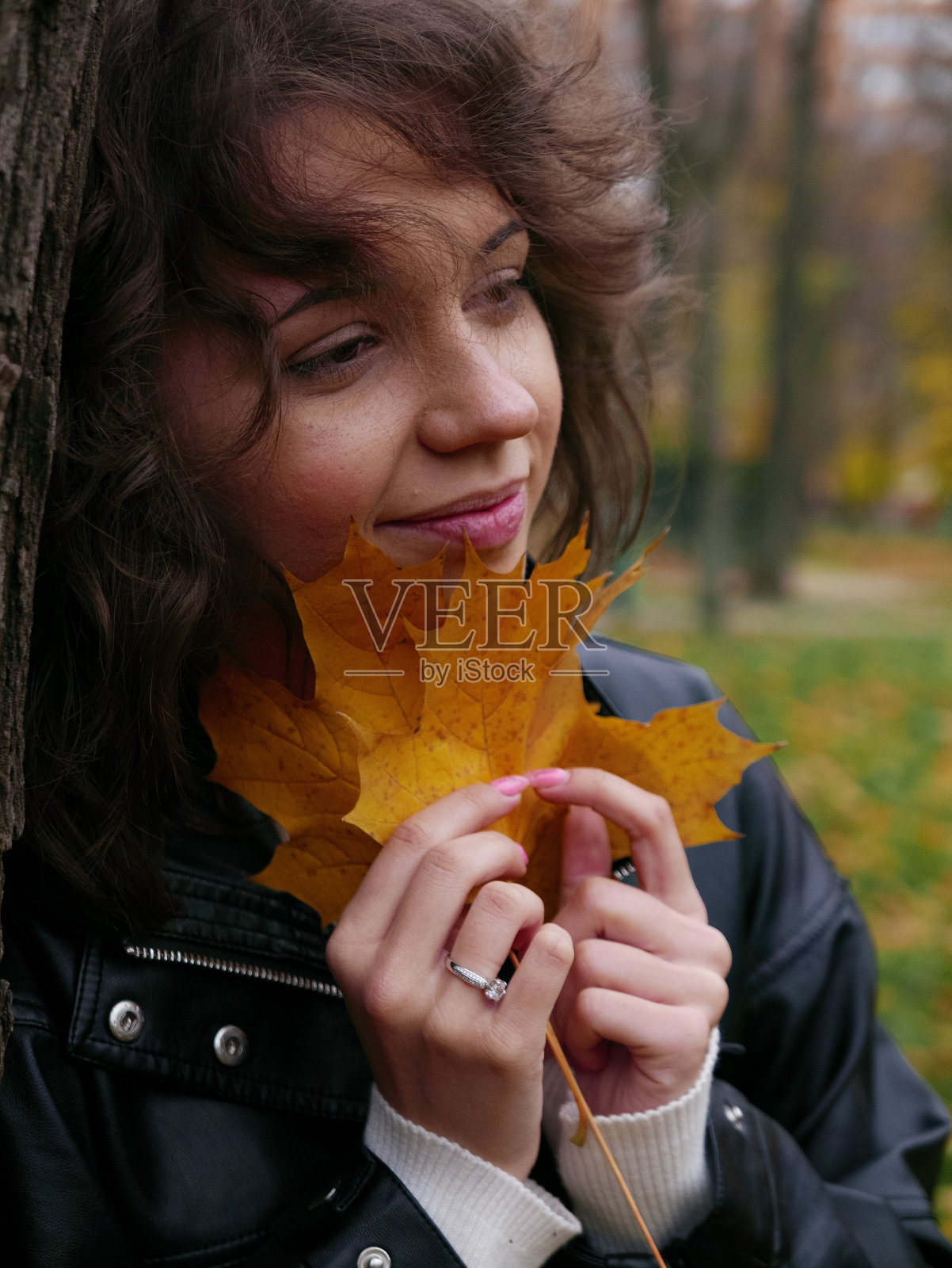 垂直的卷发黑发白人女性摆出黄色枫叶，靠在一棵树上照片摄影图片