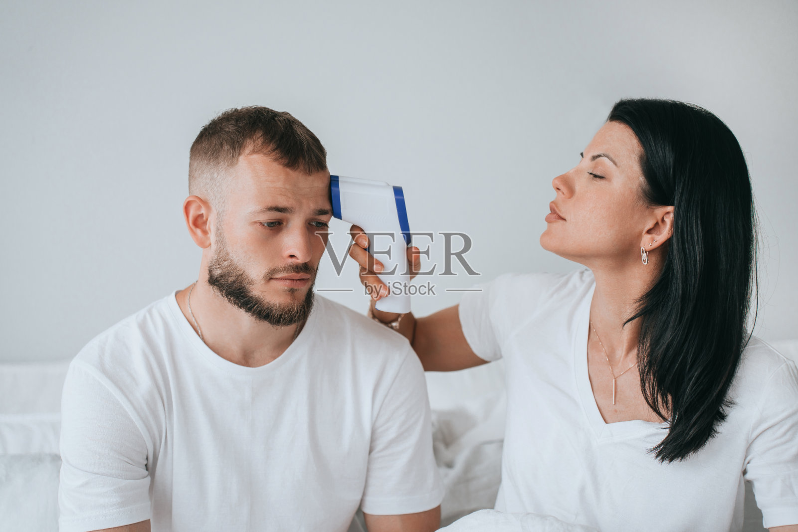 年轻的成年西班牙妇女测量她生病的丈夫坐在床上的温度。年轻的大胡子生病了，感到疲劳。意大利夫妇因为病毒呆在家里。医学治疗。照片摄影图片