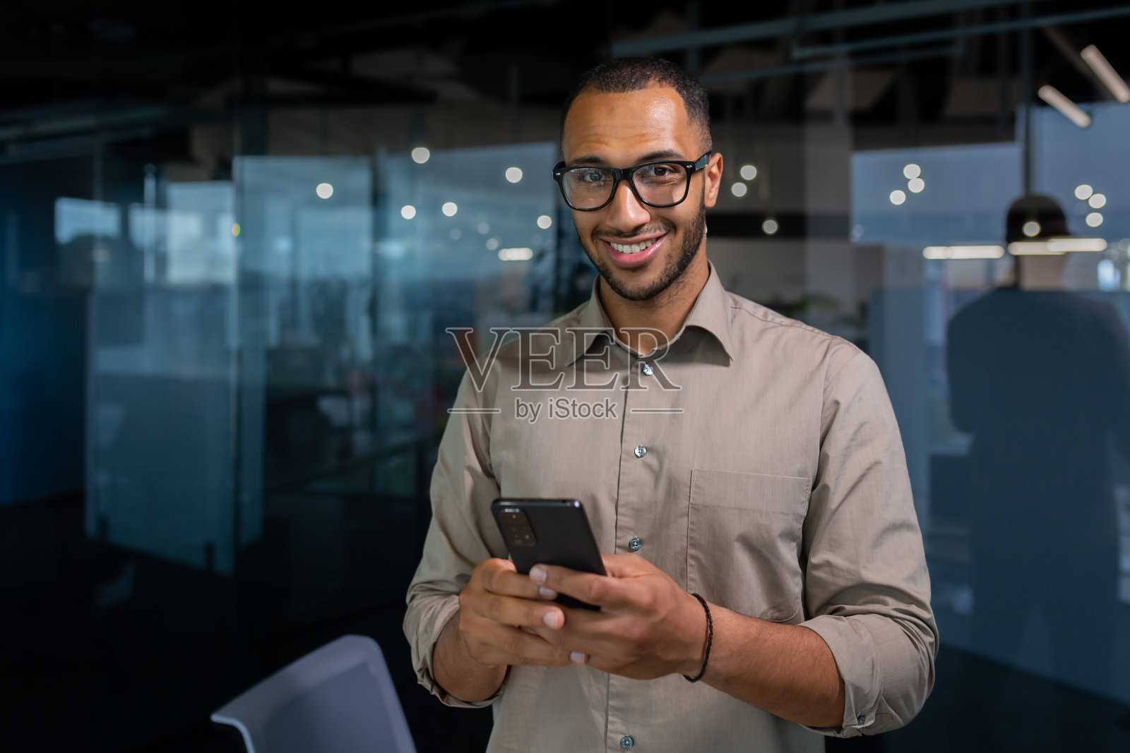 肖像年轻的西班牙裔男子在办公室里，一名穿着休闲衬衫的男子微笑着看着镜头，一名商人手里拿着智能手机，在电话上打字，打电话照片摄影图片