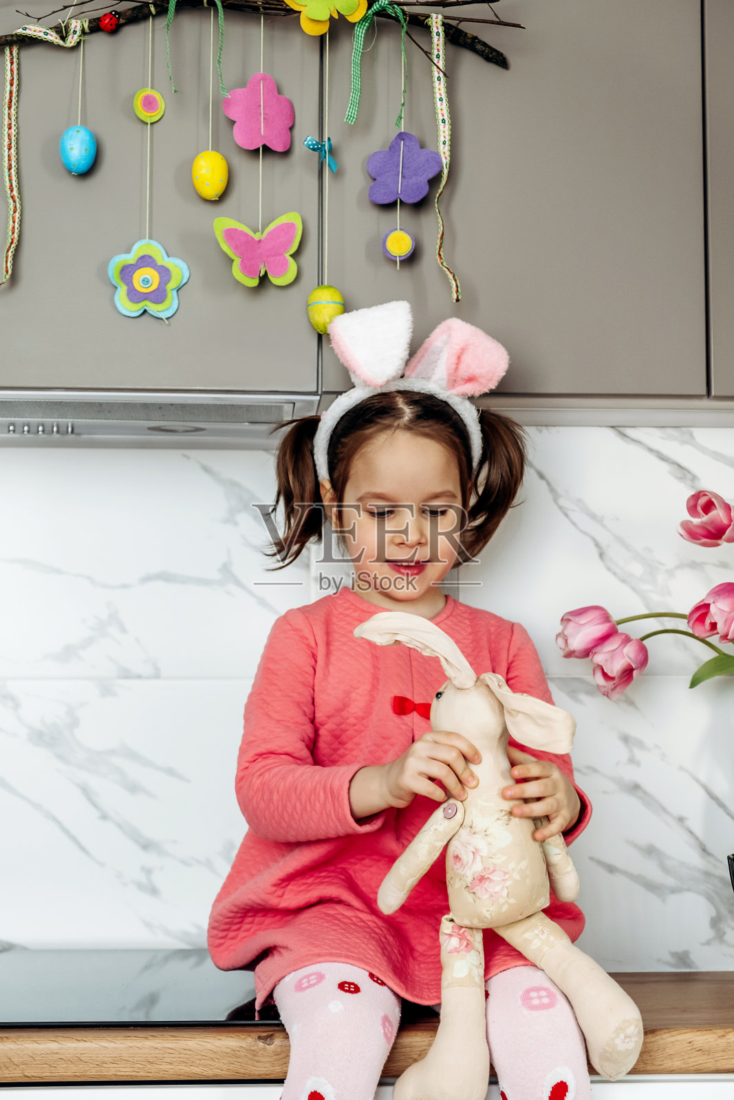 黑发小女孩穿着粉色连衣裙，头戴兔子耳朵，在厨房的台面上与玩具兔子玩耍，复活节快乐照片摄影图片