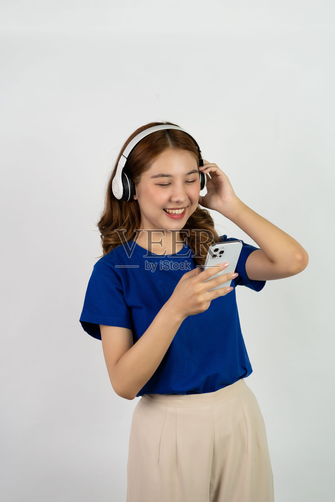 美丽的年轻女子拿着手机，戴着耳机，听着音乐，脸上带着微笑，白色背景。在线娱乐与音乐流媒体的概念照片摄影图片