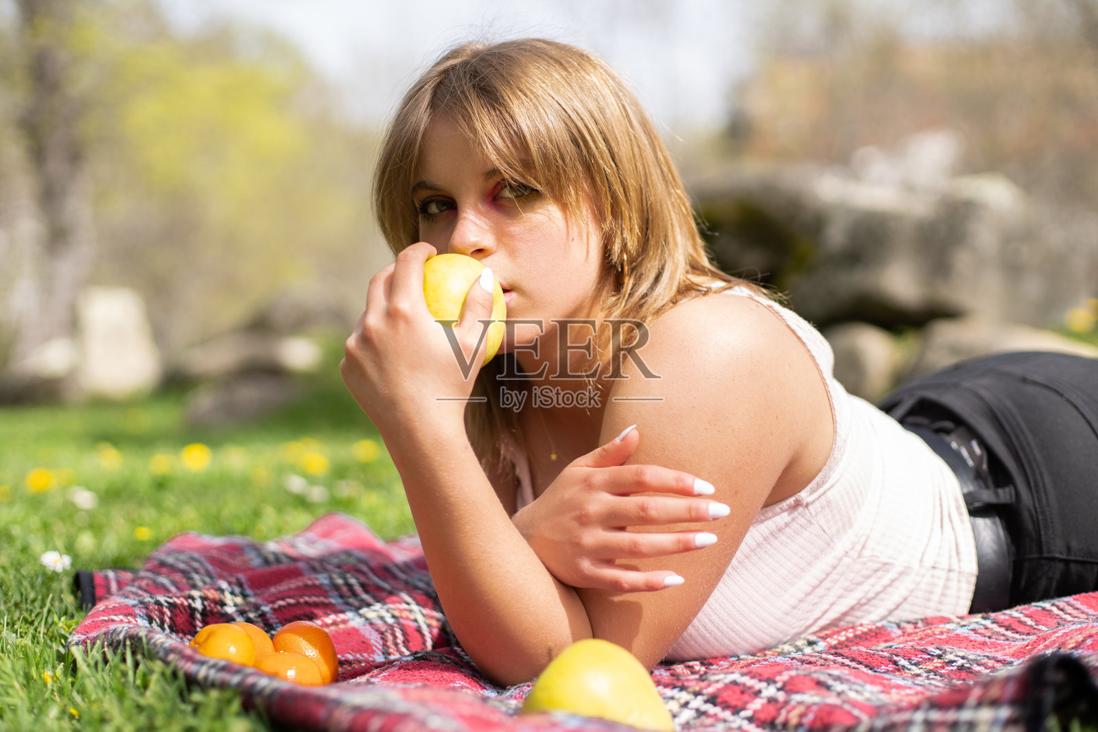 迷人的金发女人躺在草地上野餐毯在公园咬苹果照片摄影图片