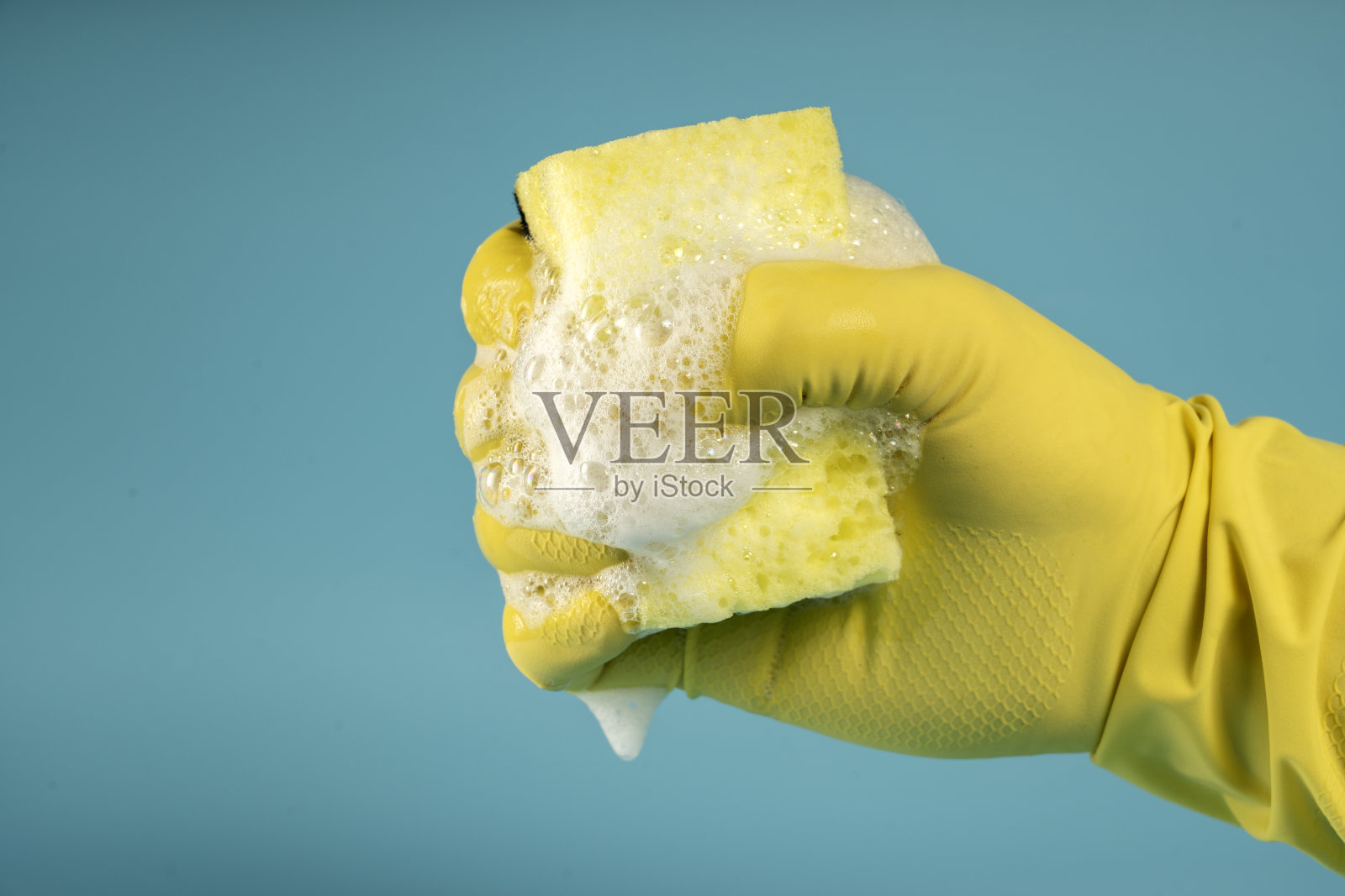 戴着黄色橡胶手套的手拿着洗碗和清洁的海绵，在蓝色背景上握紧海绵成拳头照片摄影图片