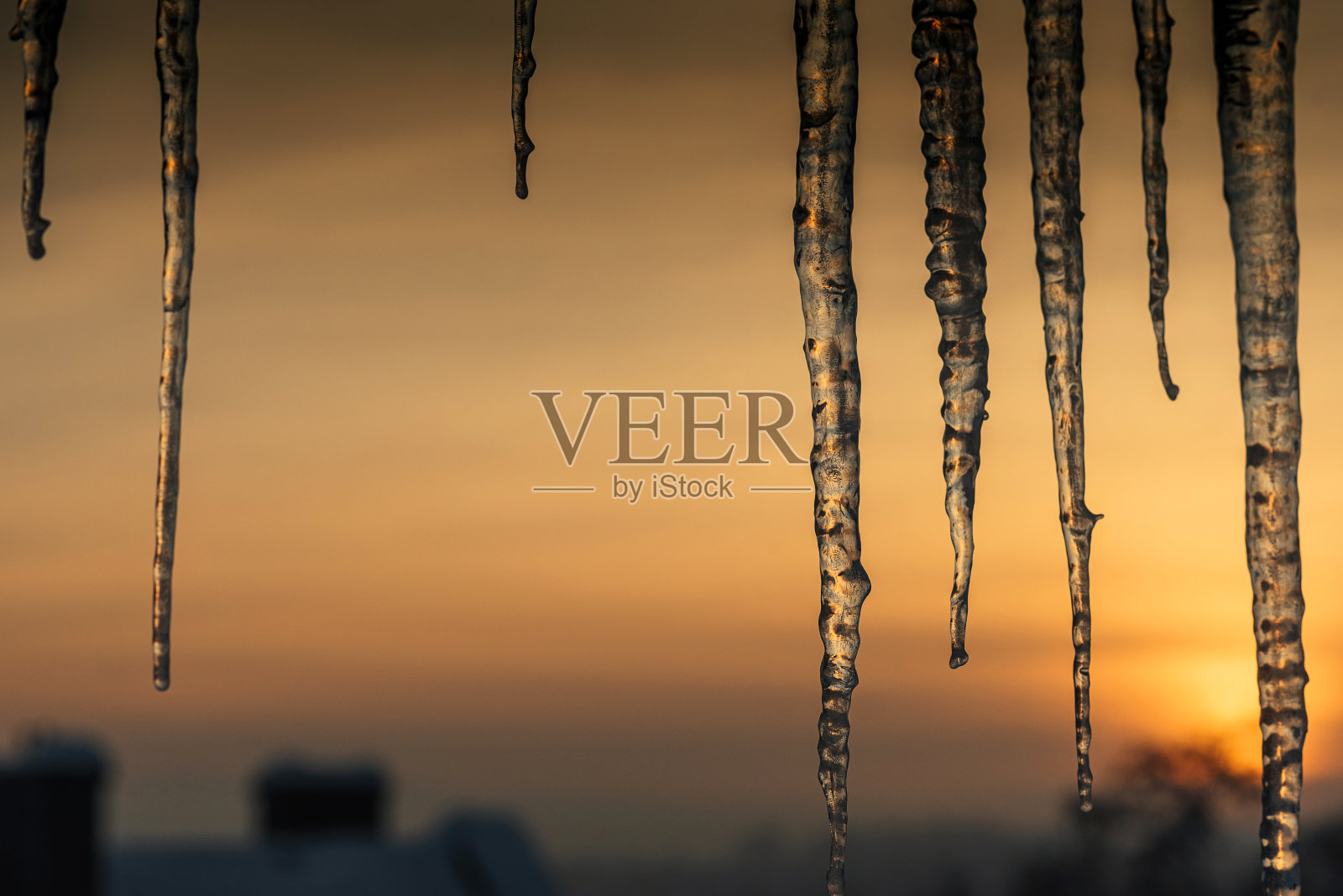 美丽的长冰柱悬挂在屋顶上，背景是金色的日落照片摄影图片