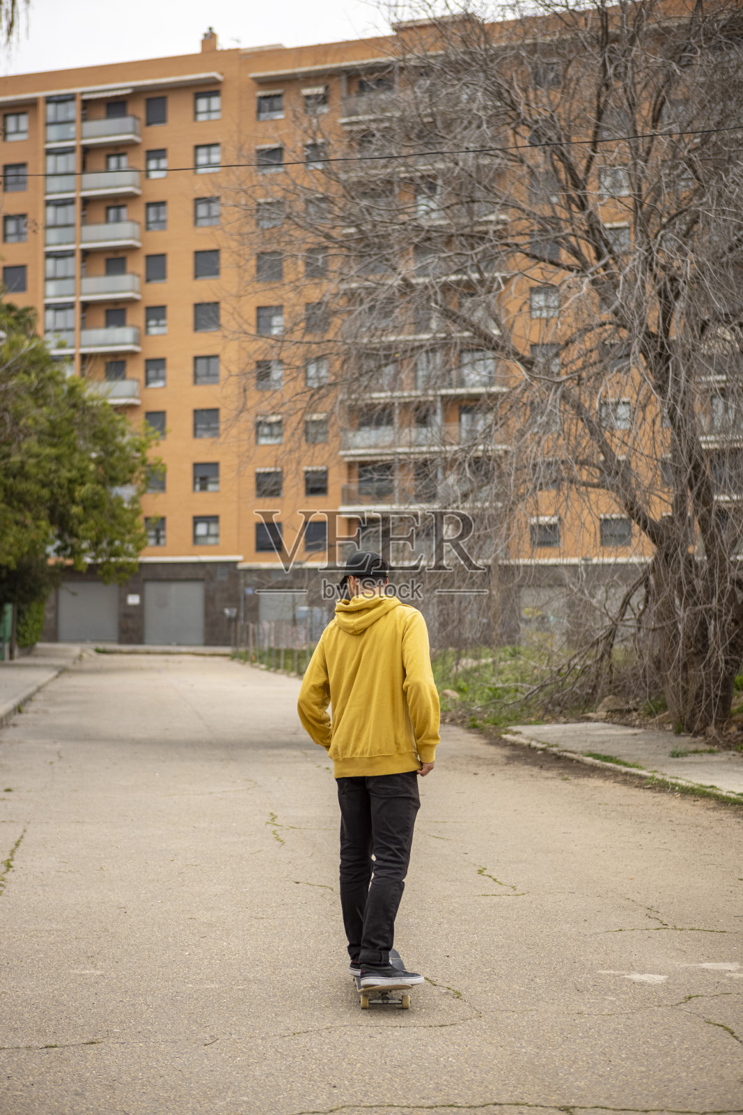 一名穿着黄色卫衣和黑色帽子的男子在一幢高层建筑的背景下踩着滑板照片摄影图片