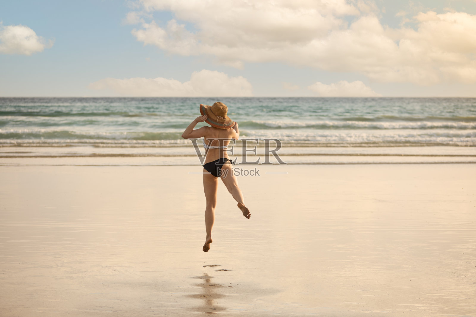 一个穿着比基尼戴着草帽的年轻女孩在海滩上跳跃照片摄影图片