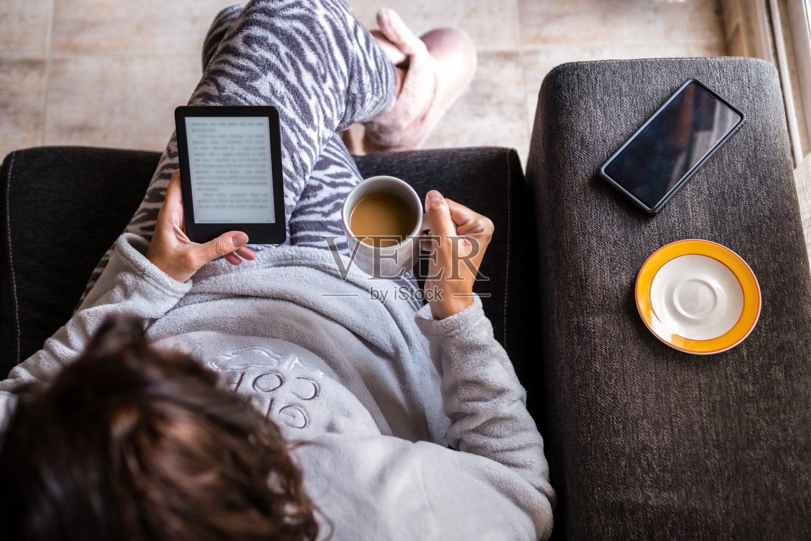 一名穿着睡衣的成年女子坐在家里客厅的沙发上，用电子书设备读着书，喝着咖啡。照片摄影图片